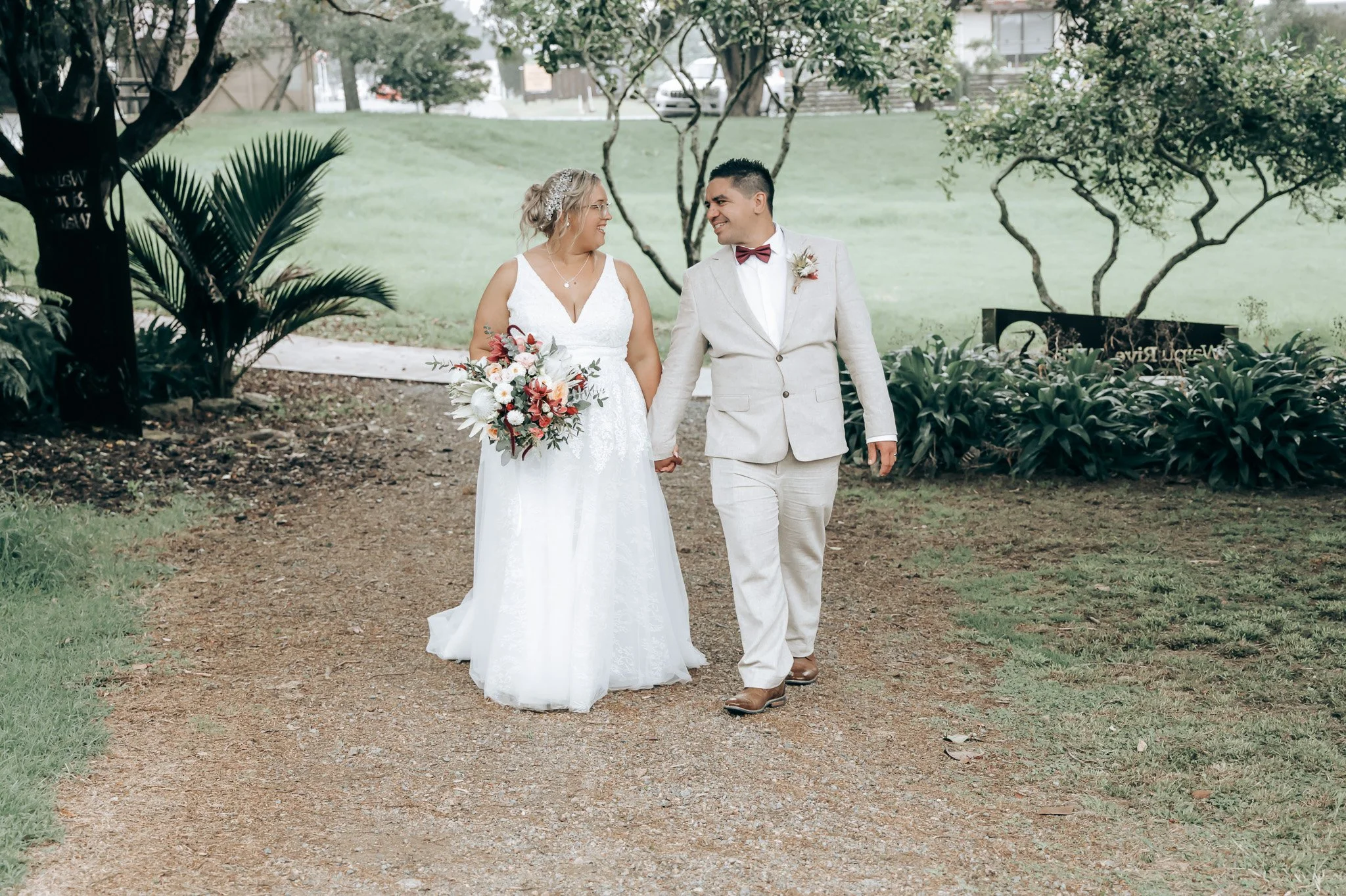 A bride and groom holding hands and walking together outdoors in a park, smiling at each other during their wedding day. The bride wears a white lace wedding dress, holding a bouquet of flowers, and has blonde hair with a decorative hairpiece. The gr