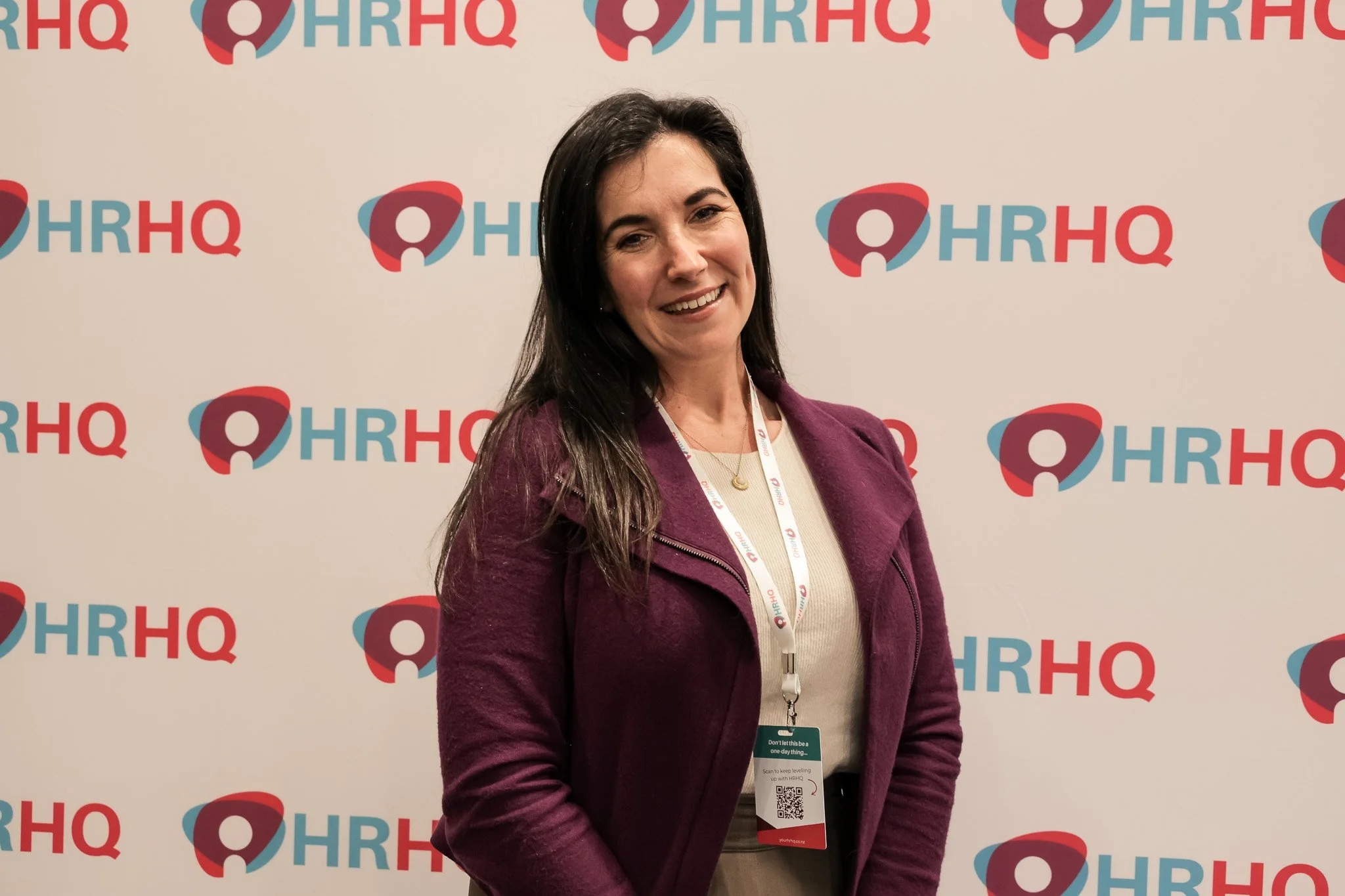 A woman with long dark hair, smiling, in front of a backdrop with the HRHQ logo, wearing a beige top and a purple jacket, with conference badge.