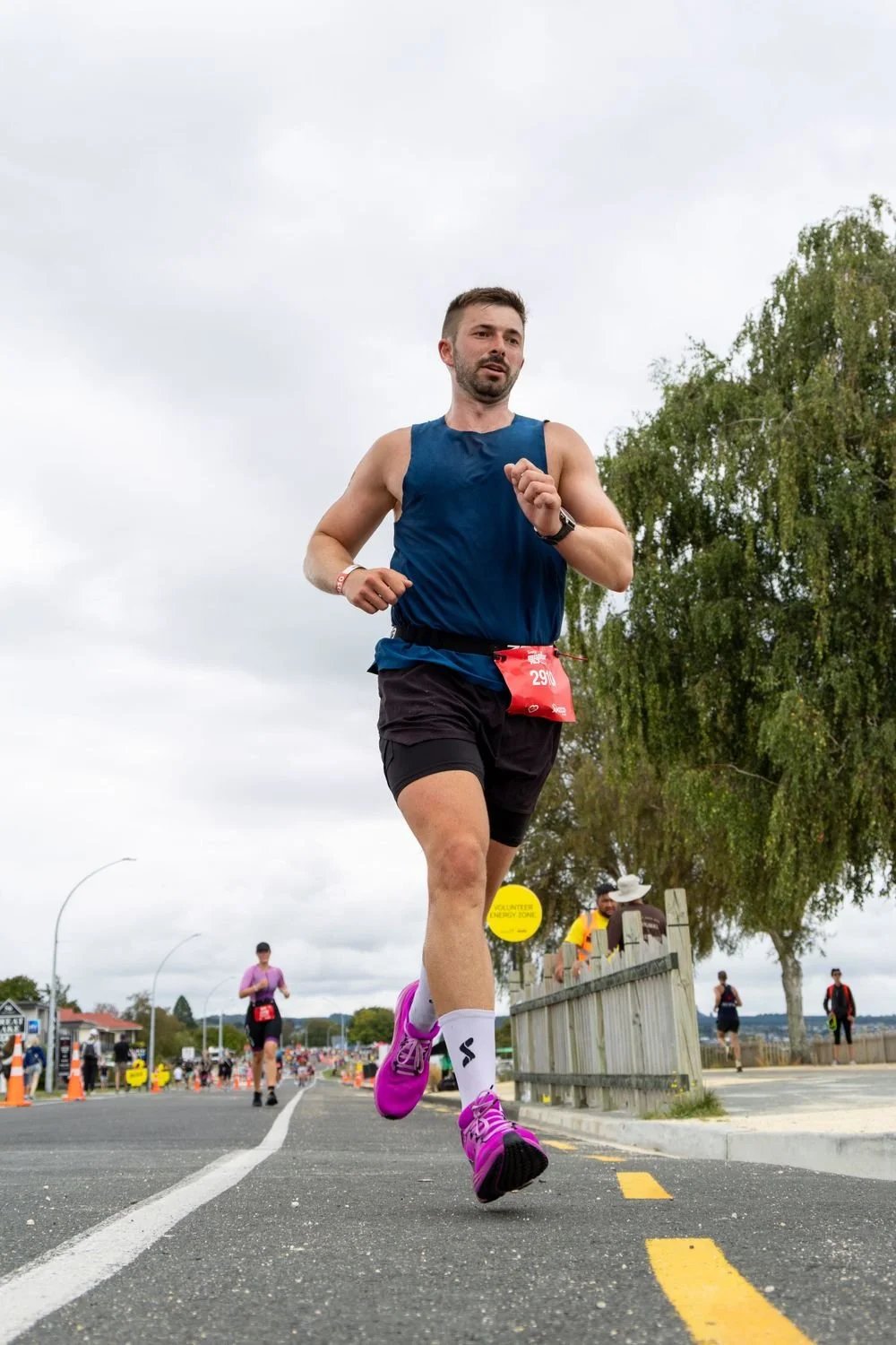 A man running in a race, wearing a blue tank top, black shorts, and pink running shoes, with other runners and spectators in the background on a cloudy day.
