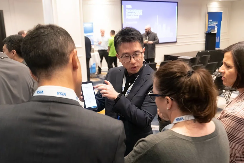 Group of five professionals in a conference room, engaged in discussion with a man showing information on a smartphone. They are wearing conference badges, and there are other attendees and a presentation screen in the background.