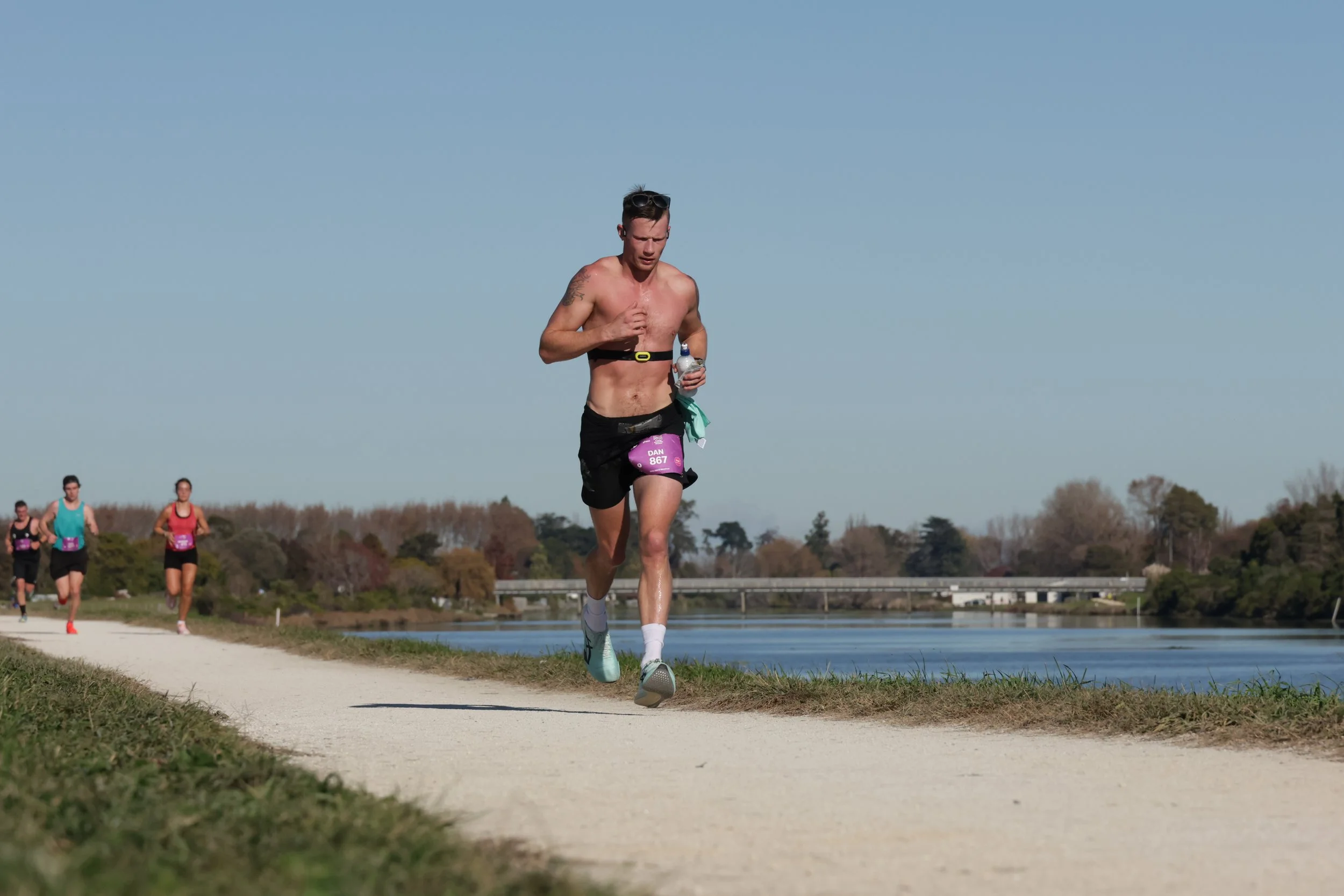 A man running shirtless on a trail near water with other runners in the background under a clear blue sky.