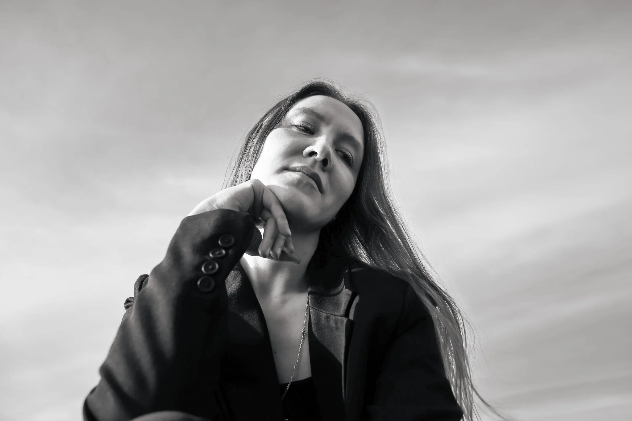 Black and white photo of a woman with long hair looking down at the camera, wearing a dark blazer and shirt, with a cloudy sky background.