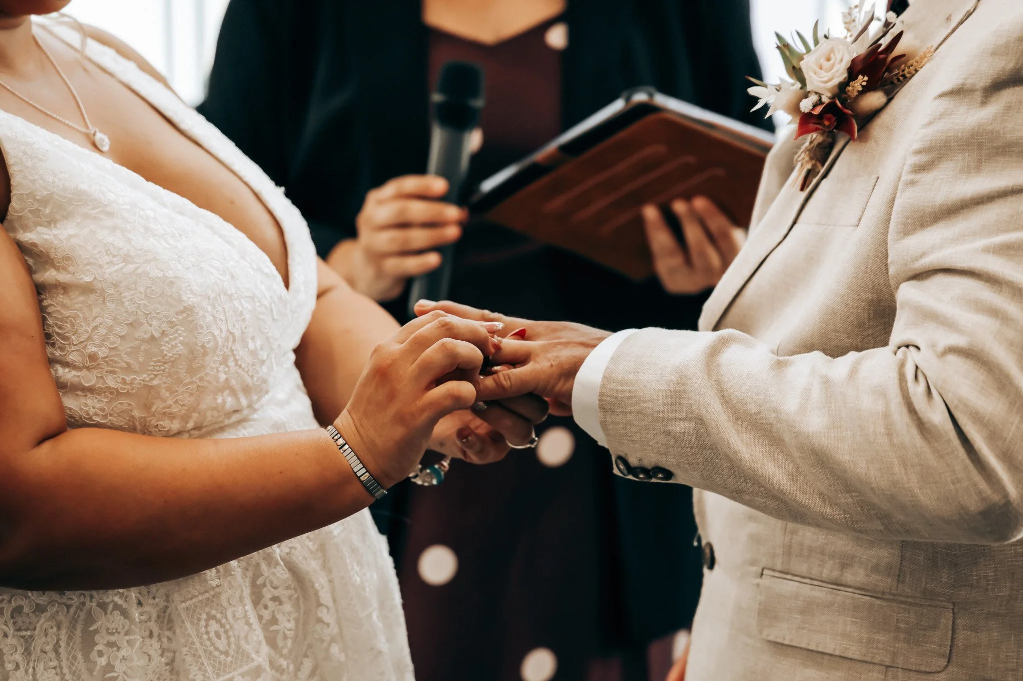 A bride and groom exchanging wedding rings during their ceremony, with a person officiating in the background holding a microphone and a folder, close-up of their hands, the bride in a lace wedding dress and the groom in a light-colored suit with a b