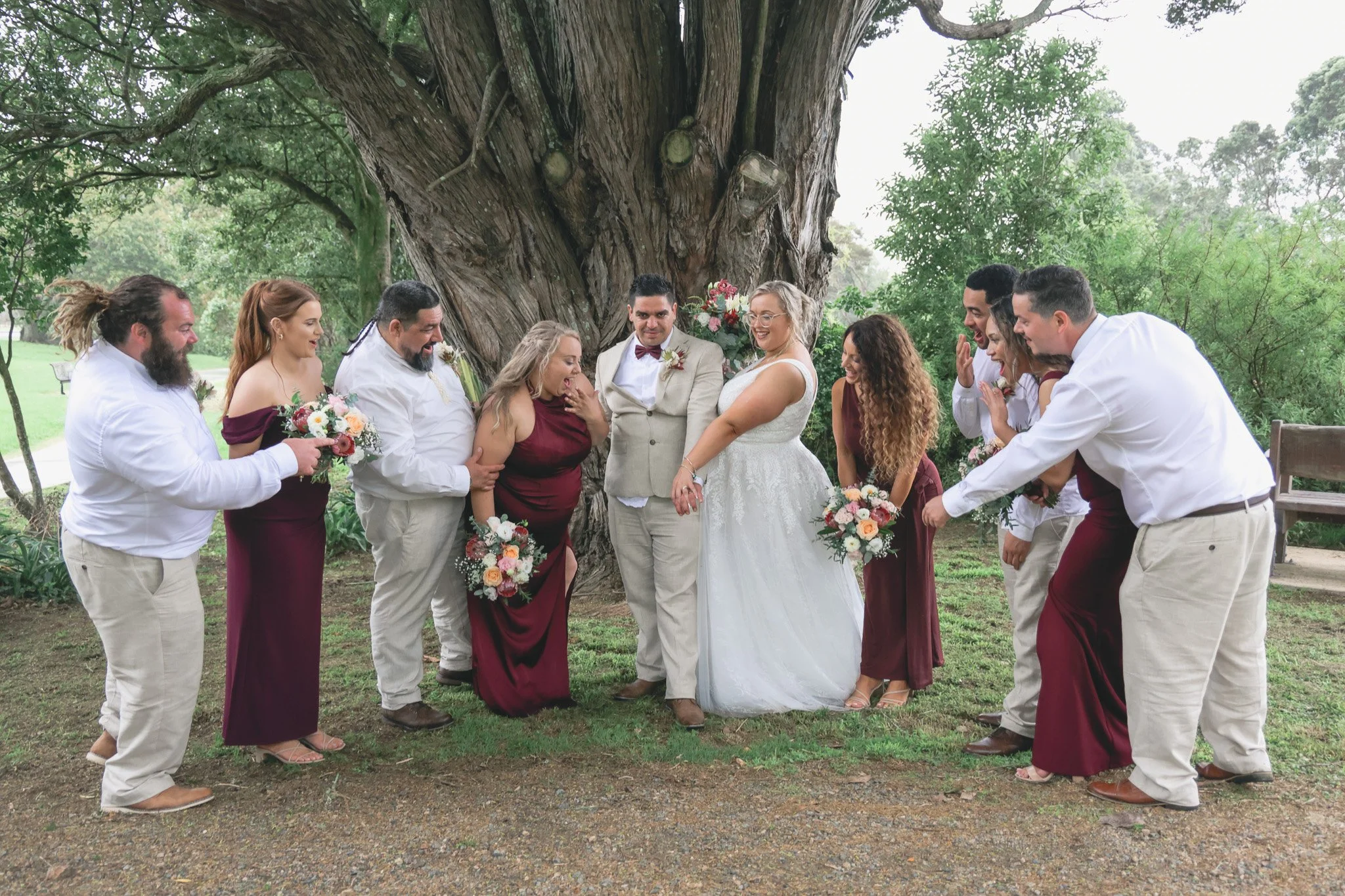 A wedding ceremony taking place outdoors beneath a large tree, with the bride, dressed in a white gown, and the groom, wearing a beige suit, surrounded by bridesmaids and groomsmen in formal attire, holding bouquets of flowers.