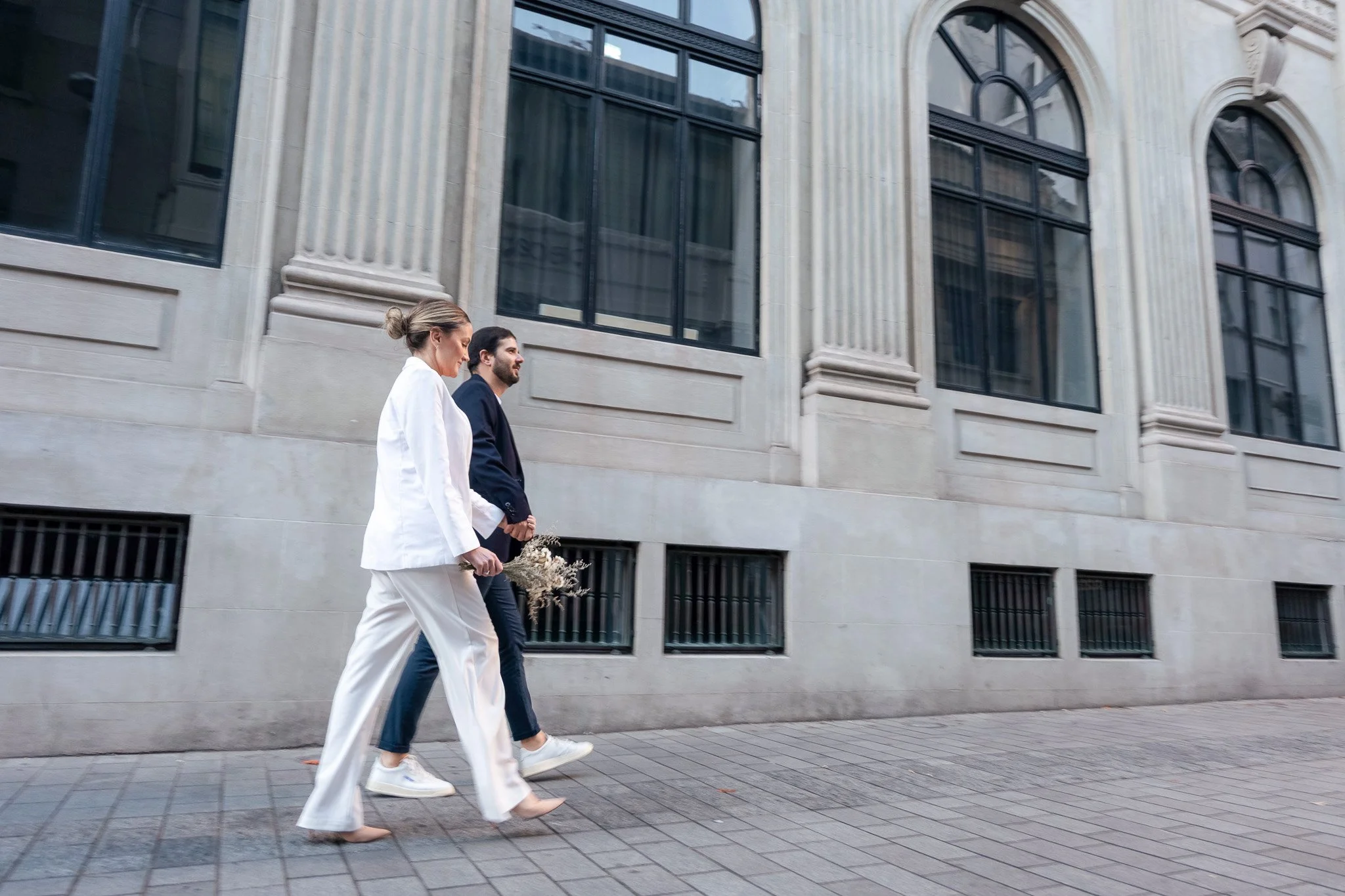 A couple walking on a city sidewalk in front of a large building with arched windows, the woman in a white outfit holding a small bouquet of dried flowers.