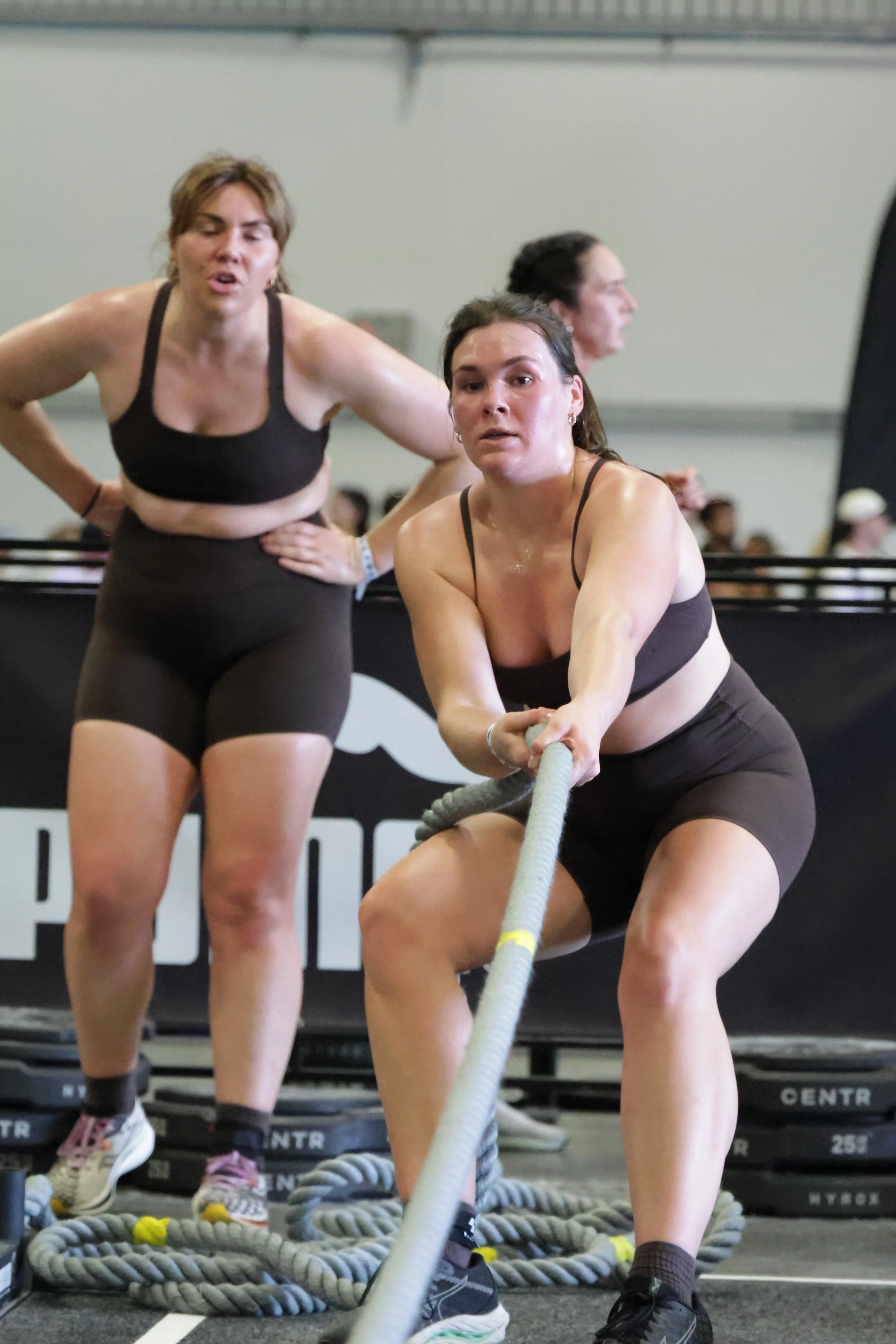 Two women participating in a strongwoman competition, pulling a weighted rope in a gym setting. A woman with dark hair and a black sports bra is in the foreground, and another woman with short hair and a black workout top is in the background. There 