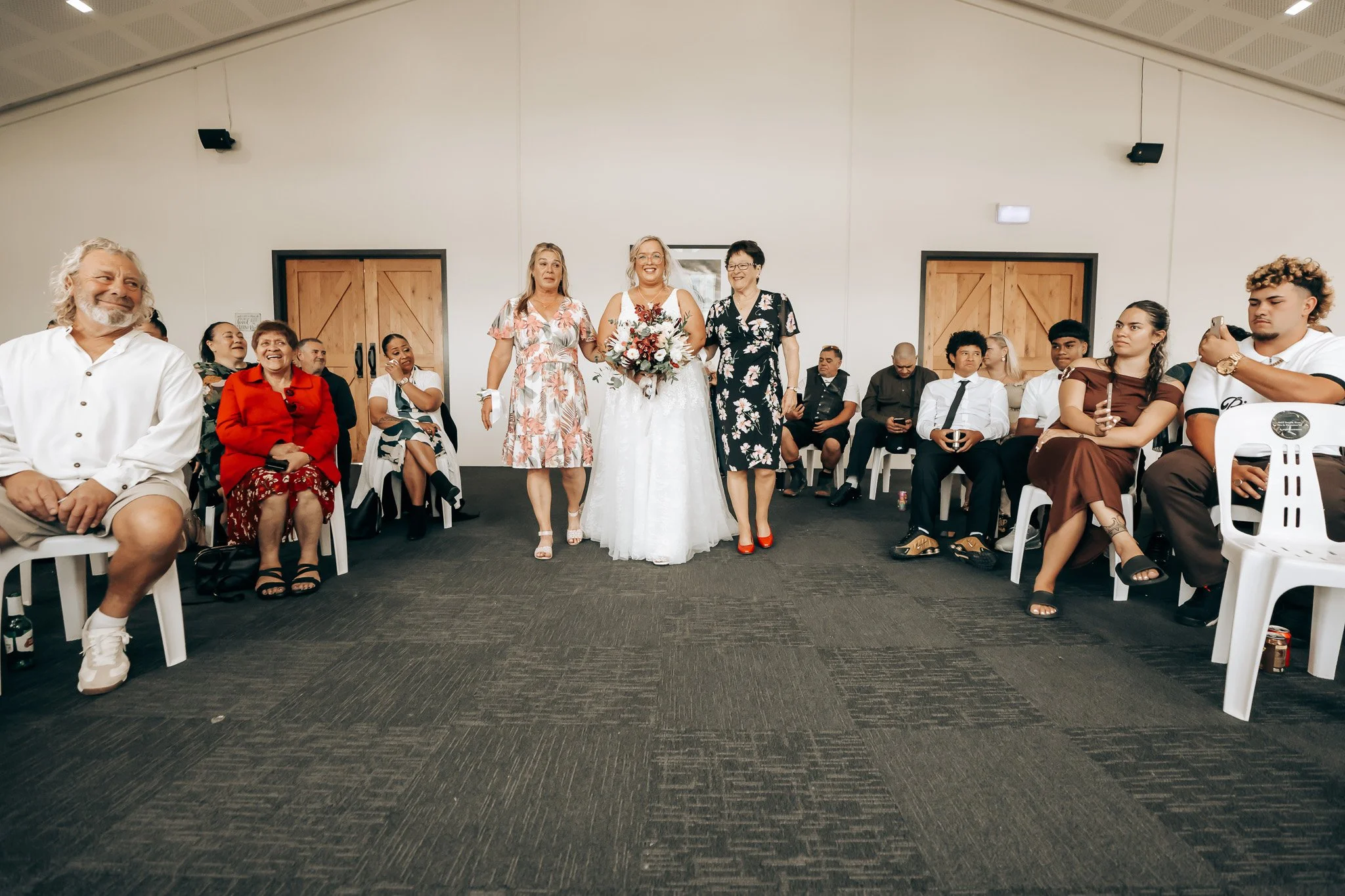 A group of people at a wedding, with three women at the center, one in a wedding dress holding a bouquet, flanked by two women in floral dresses, all smiling. Other guests sit around, some wearing formal attire, in a room with wooden doors and a pitc