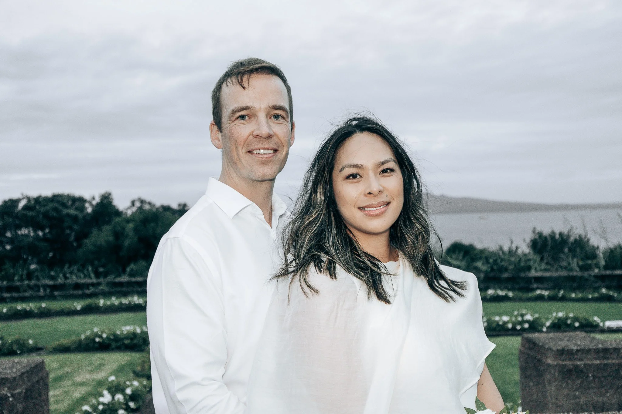 A man and a woman standing outdoors near water and trees on a cloudy day.