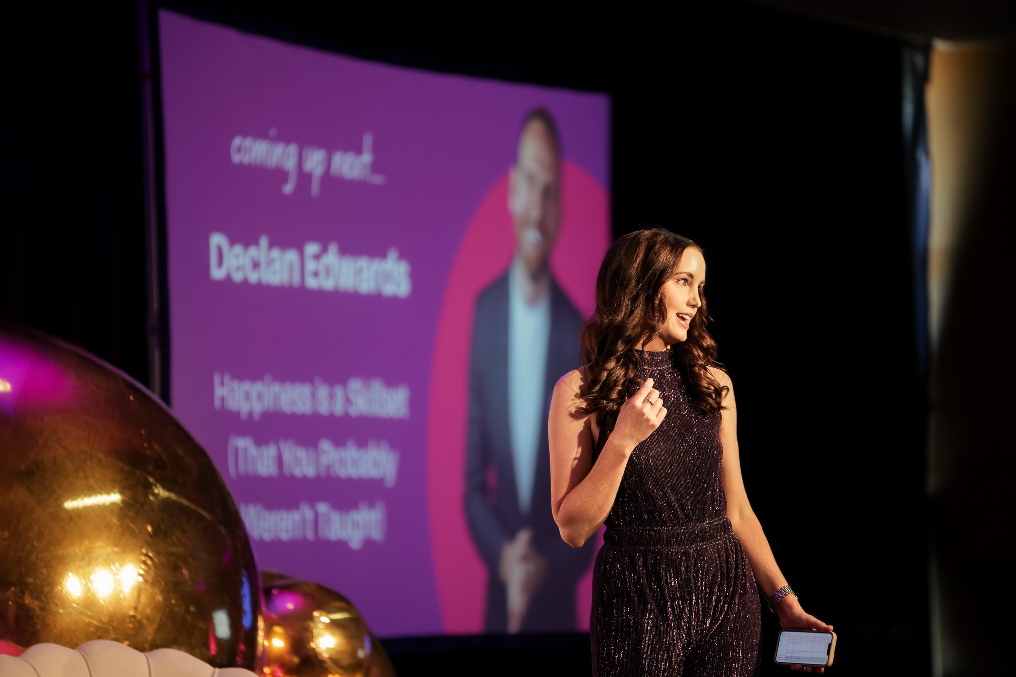 A woman in a sparkly dress is speaking at a conference or event, holding a tablet in her left hand. She is standing in front of a purple screen with text and a blurred image of a person. There are large, shiny gold balloons in the foreground.
