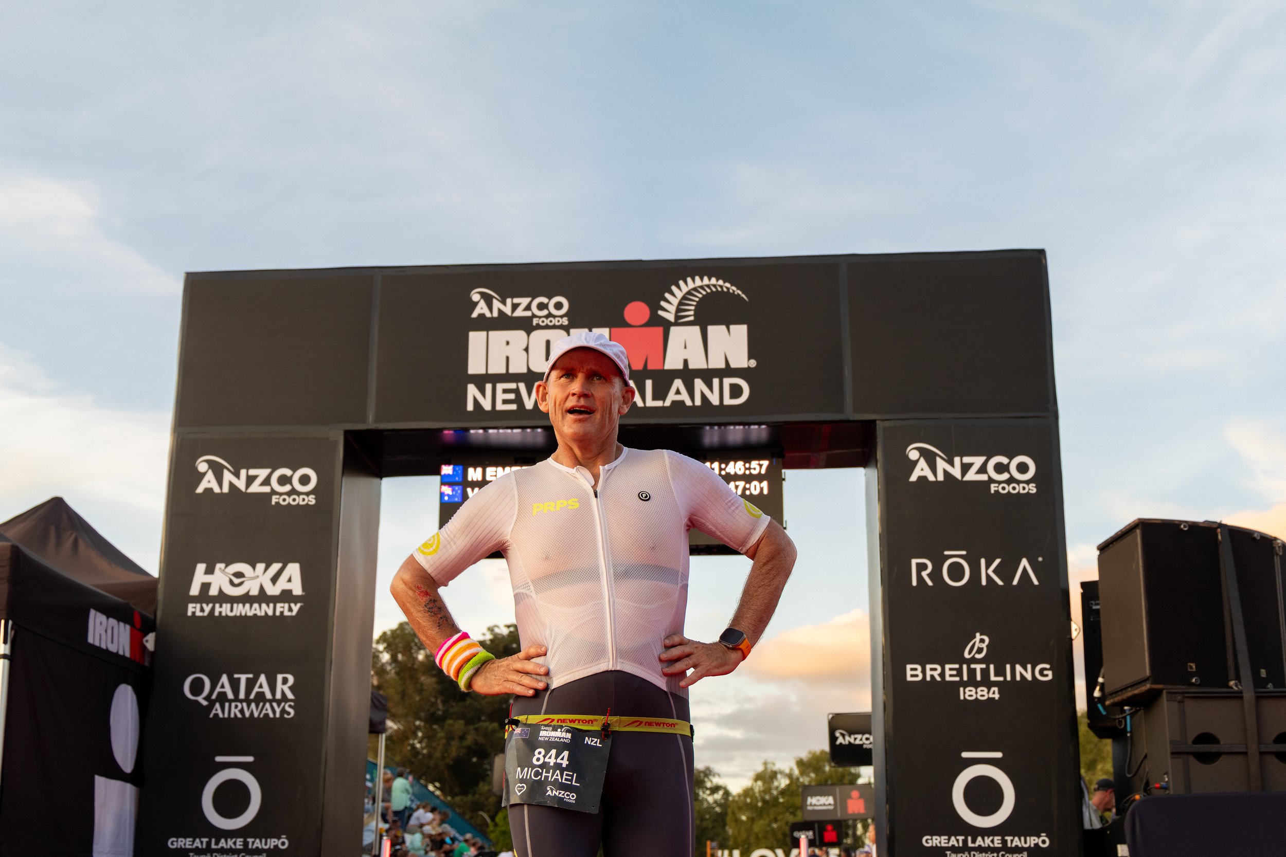 Male triathlete wearing a white cap and race gear, standing with hands on hips at the Ironman New Zealand finish line, with event banners and digital display behind and in the background.