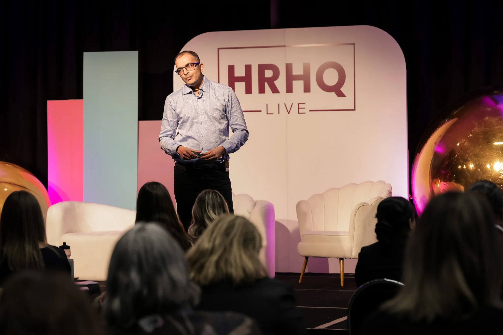 A man in glasses and a light-colored shirt stands on stage in front of an audience at HRHQ Live, with decorative chairs and large golden balloons on stage.