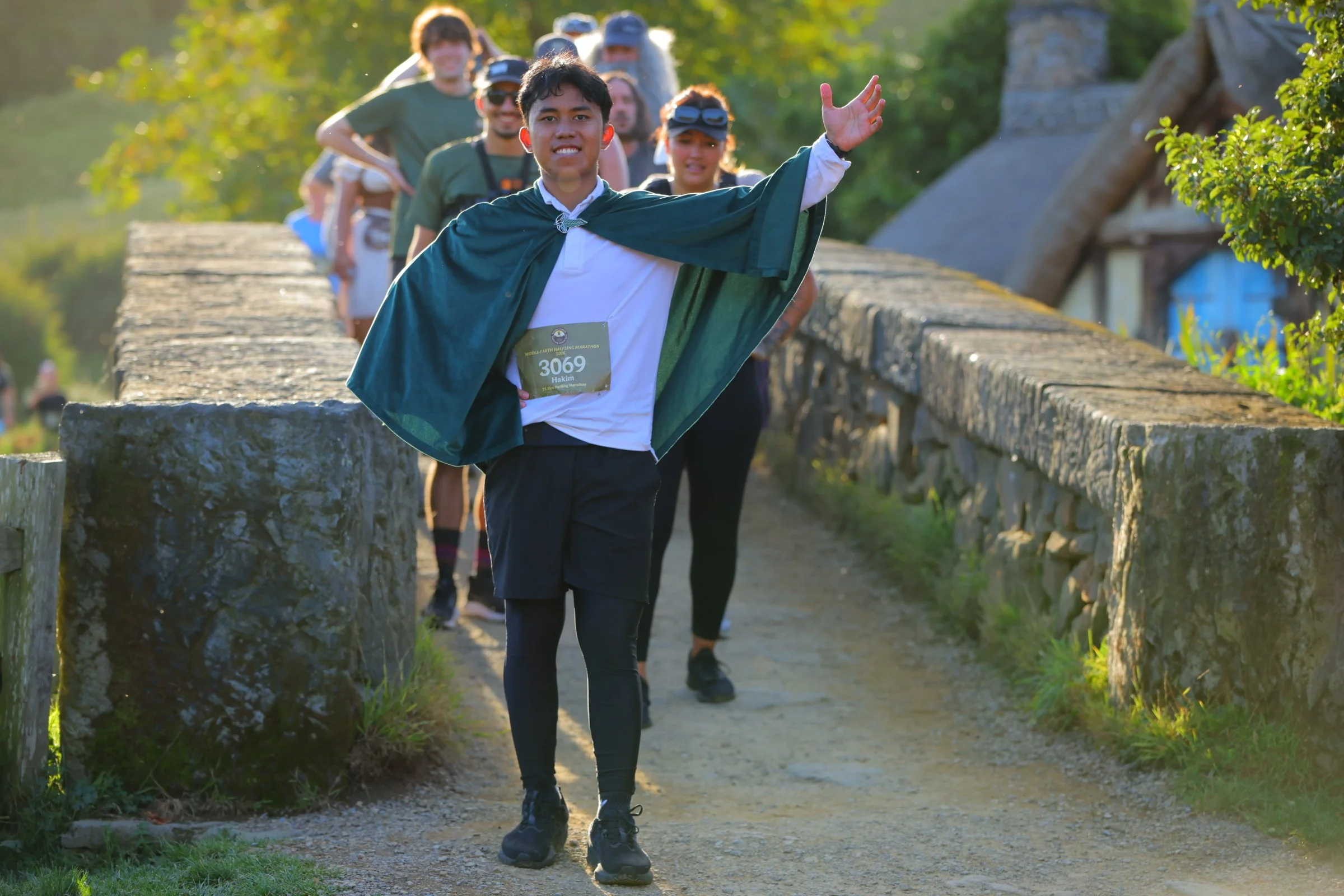 A group of people walking on a stone and dirt path outdoors, with the person in front waving and wearing a green cape over a white shirt, with a race bib. The sunlight shines through trees, creating a bright and lively atmosphere.