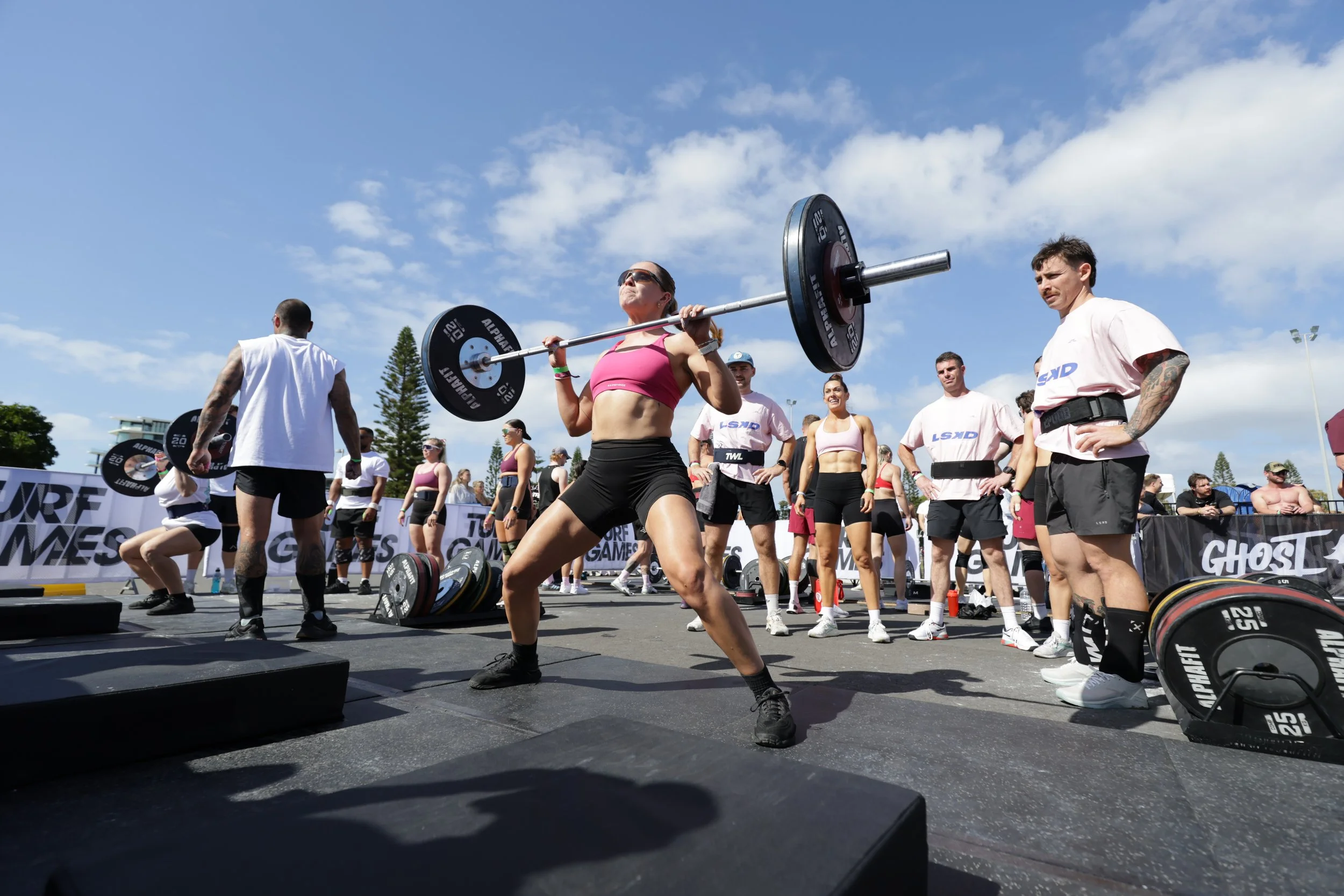 A woman in a pink sports bra and black shorts lifting a large barbell with weights at an outdoor fitness event, surrounded by other participants and spectators, under a partly cloudy sky.