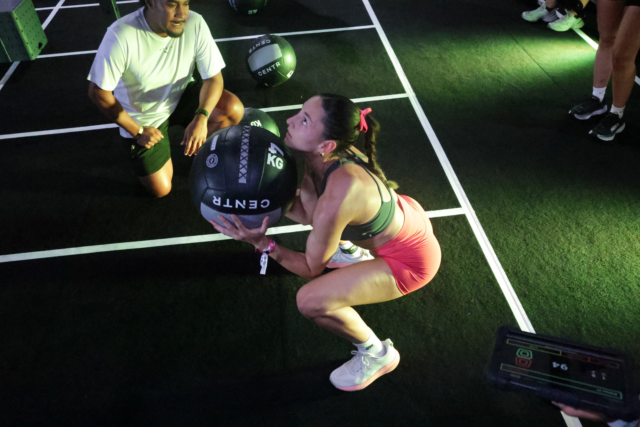 A woman in athletic clothing performing a workout exercise with a medicine ball in a gym, supervised by a trainer kneeling beside her. There are other gym members and equipment around.