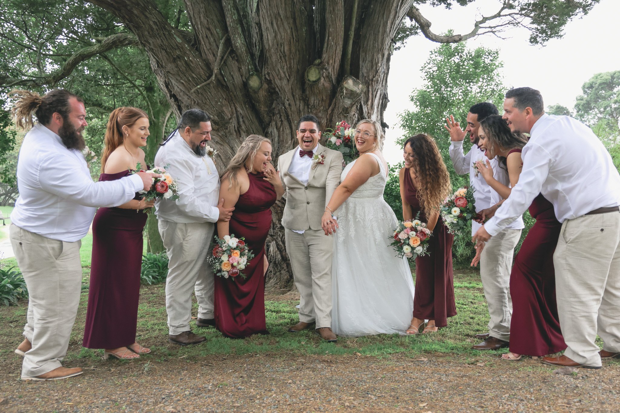 A wedding party standing outdoors in front of a large tree, with the bride and groom at the center, surrounded by friends and family dressed in coordinated attire, all smiling and laughing.