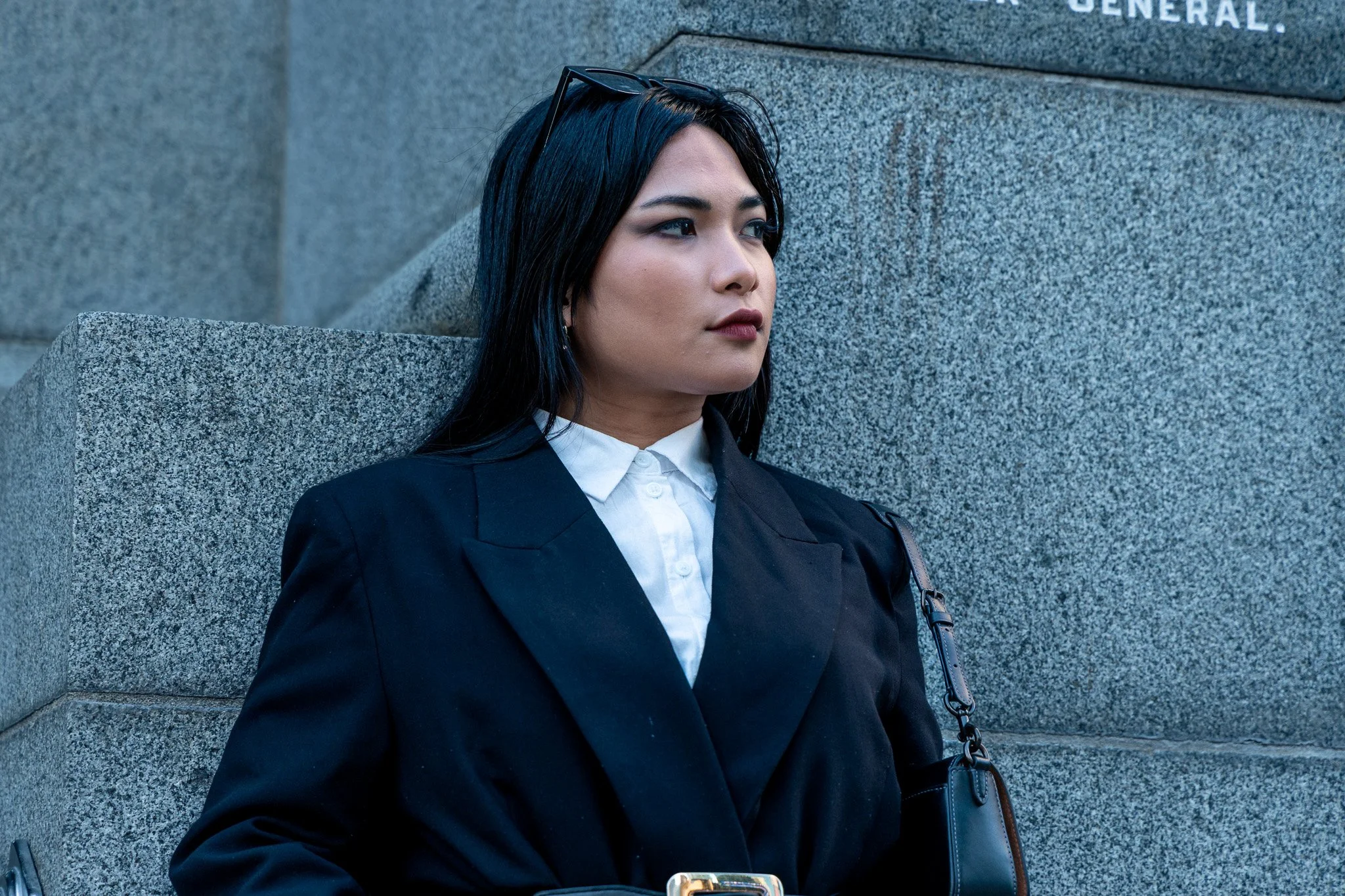A woman with dark hair and wearing a black blazer and white shirt sitting against a stone wall, with sunglasses on her head and a small black handbag hanging from her shoulder.