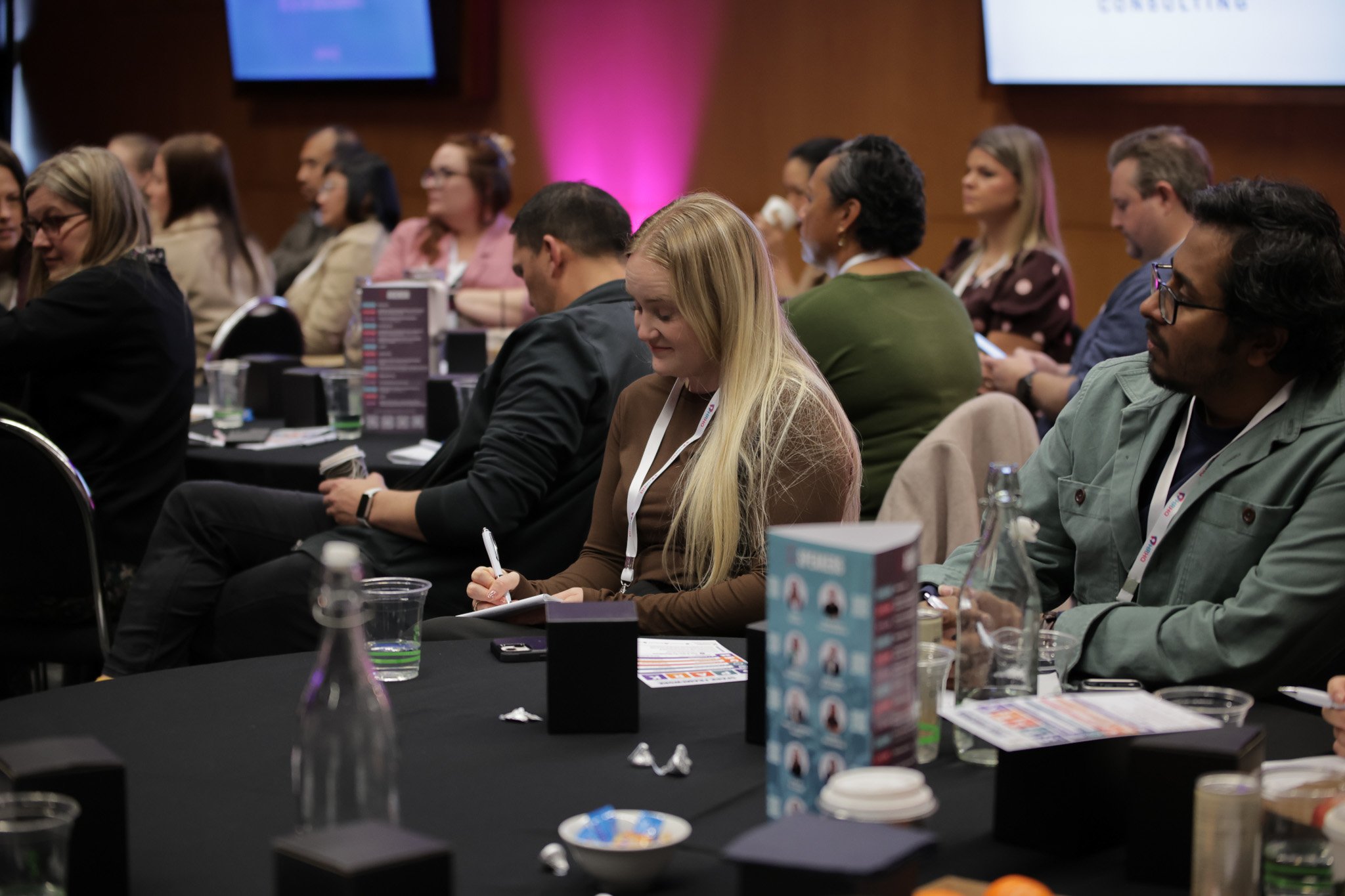 Conference attendees seated at tables in a large room, listening to a presentation, with presentation screens in the background.