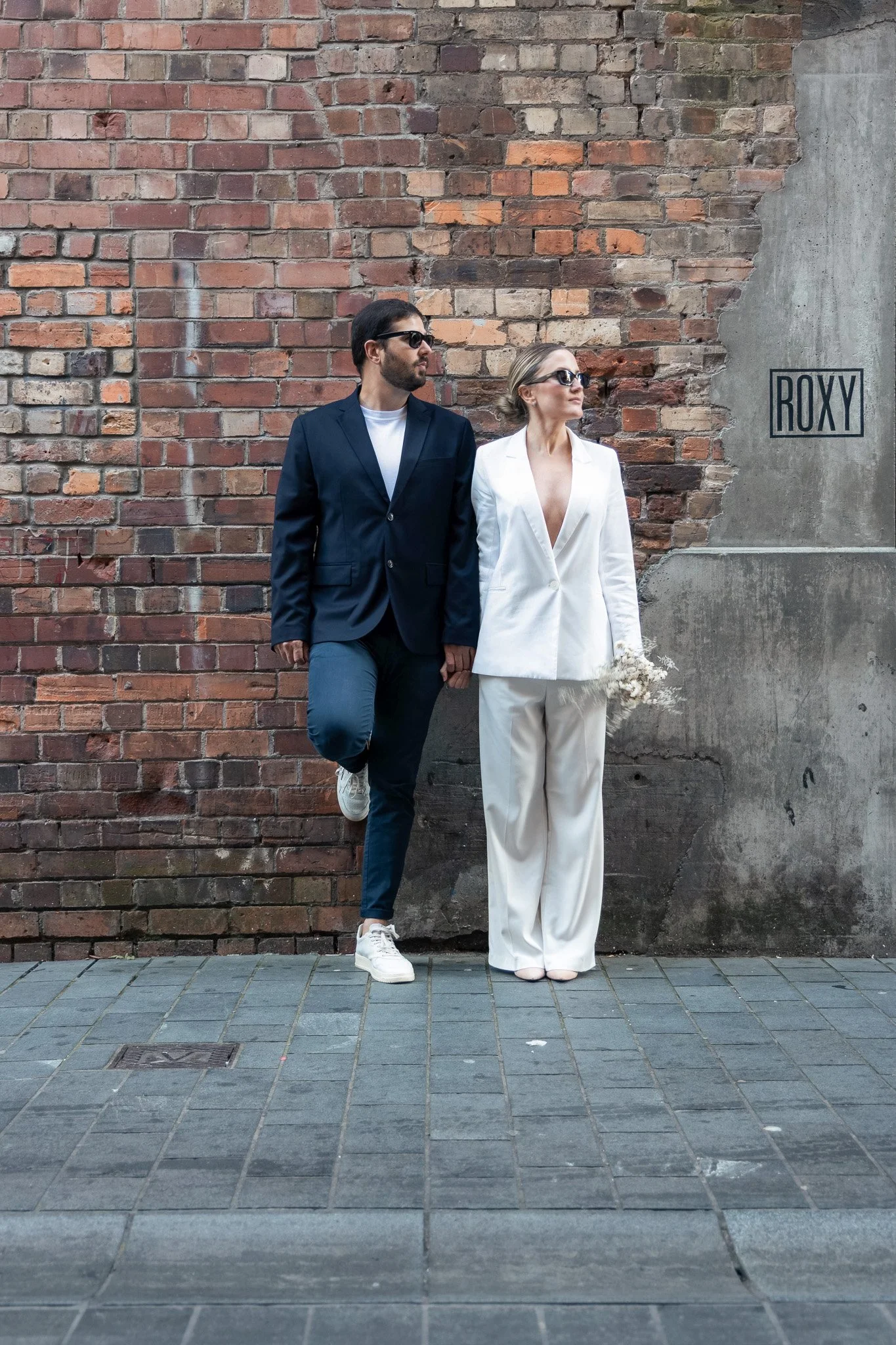 A man and woman standing against a brick wall, both wearing sunglasses, with the man in a navy blazer and white shirt, and the woman in a white suit and holding flowers, on a city sidewalk.