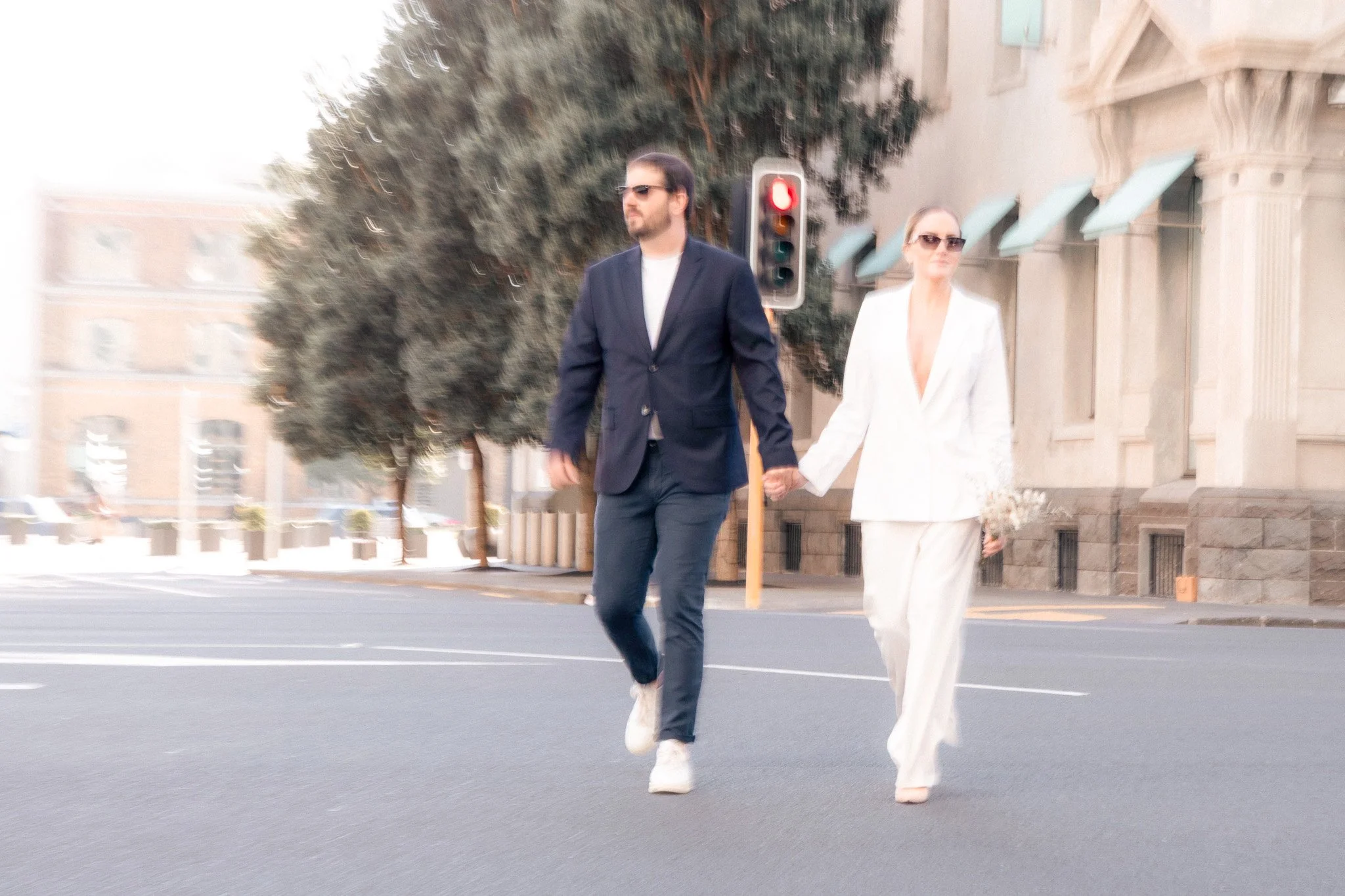 A man and woman walking hand in hand across a city street during daytime. The man is wearing a dark blazer, white shirt, sunglasses, and jeans. The woman is wearing a white blazer and pants, and sunglasses. There is a tree, a traffic light showing re