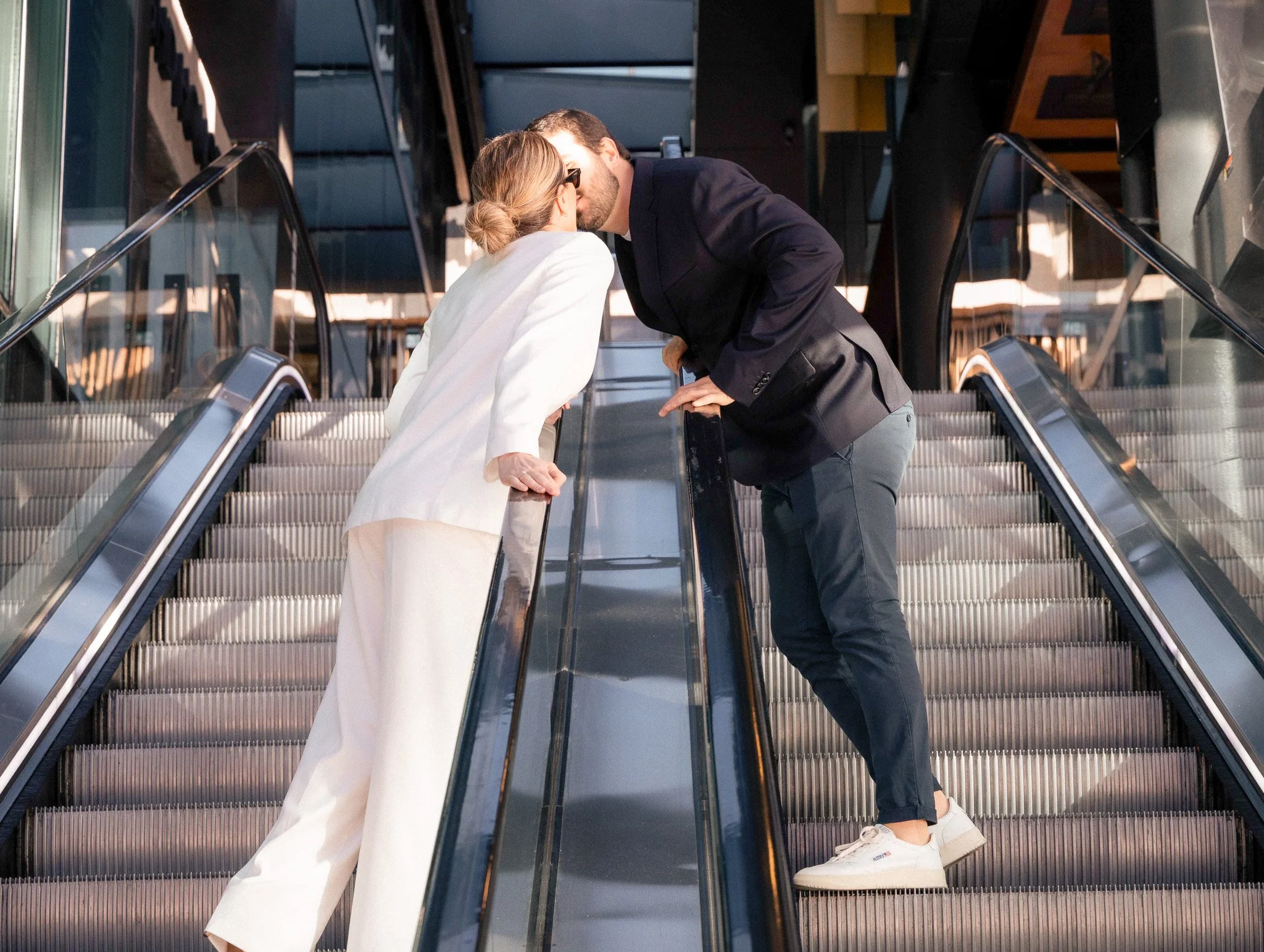 A man in a black blazer and grey pants leaning down to kiss a woman in a white suit and glasses on an escalator in a shopping mall.