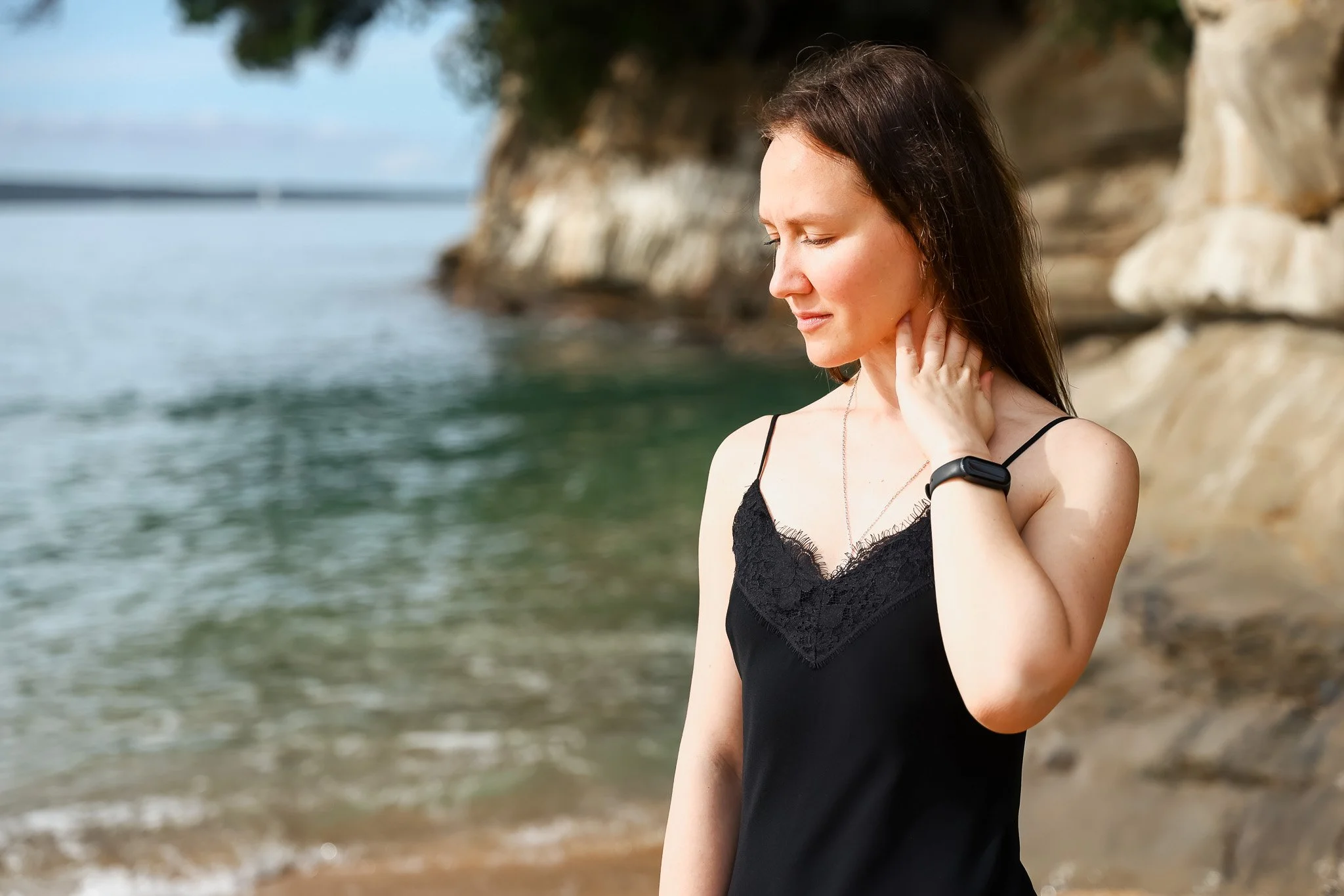 A young woman standing on a beach with her eyes closed, touching her neck, wearing a black sundress with lace details and a black bracelet, near rocky cliffs and water.