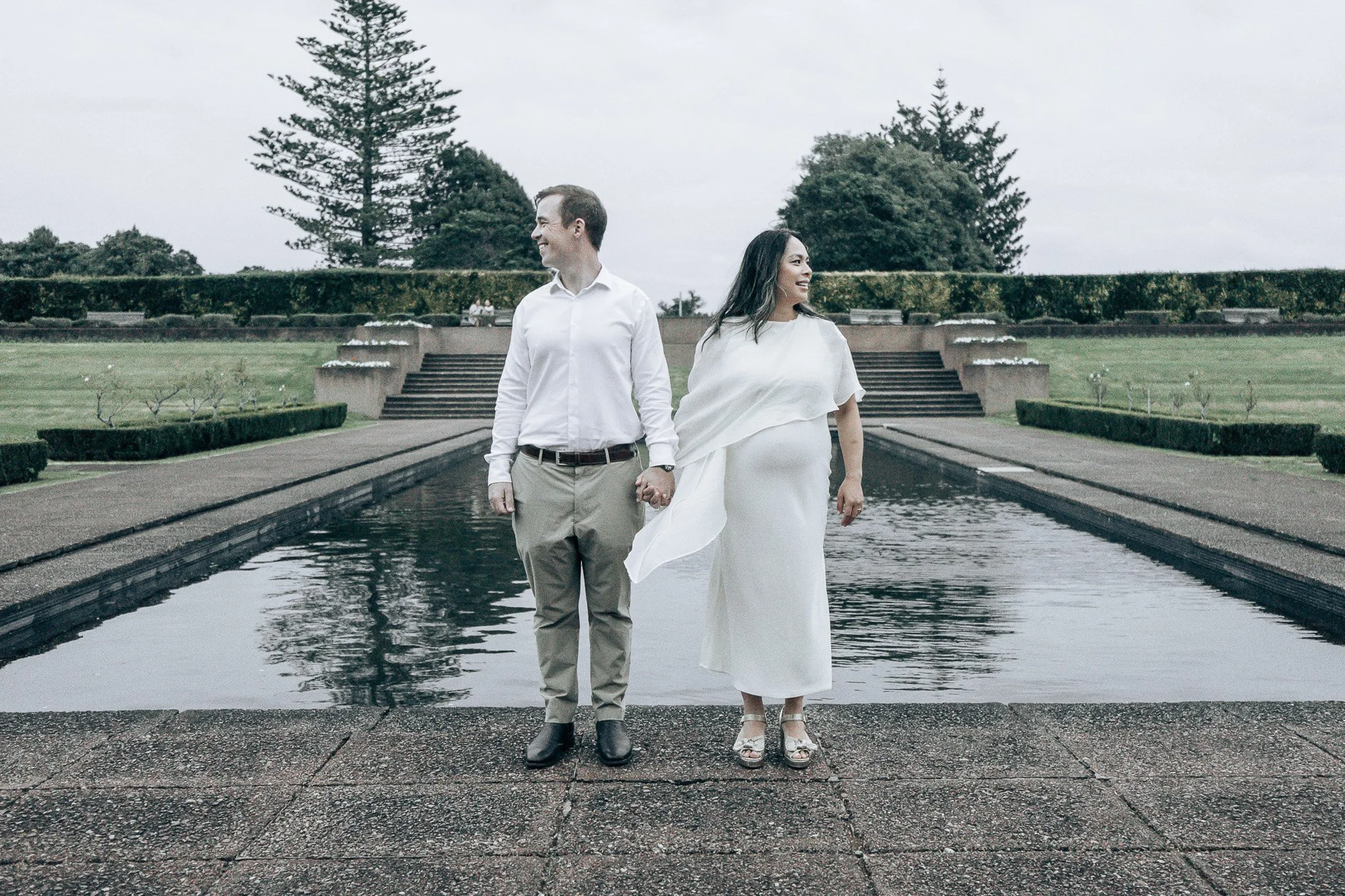 A smiling man and pregnant woman holding hands, walking together by a reflective water feature in a park with trees and concrete steps in the background.