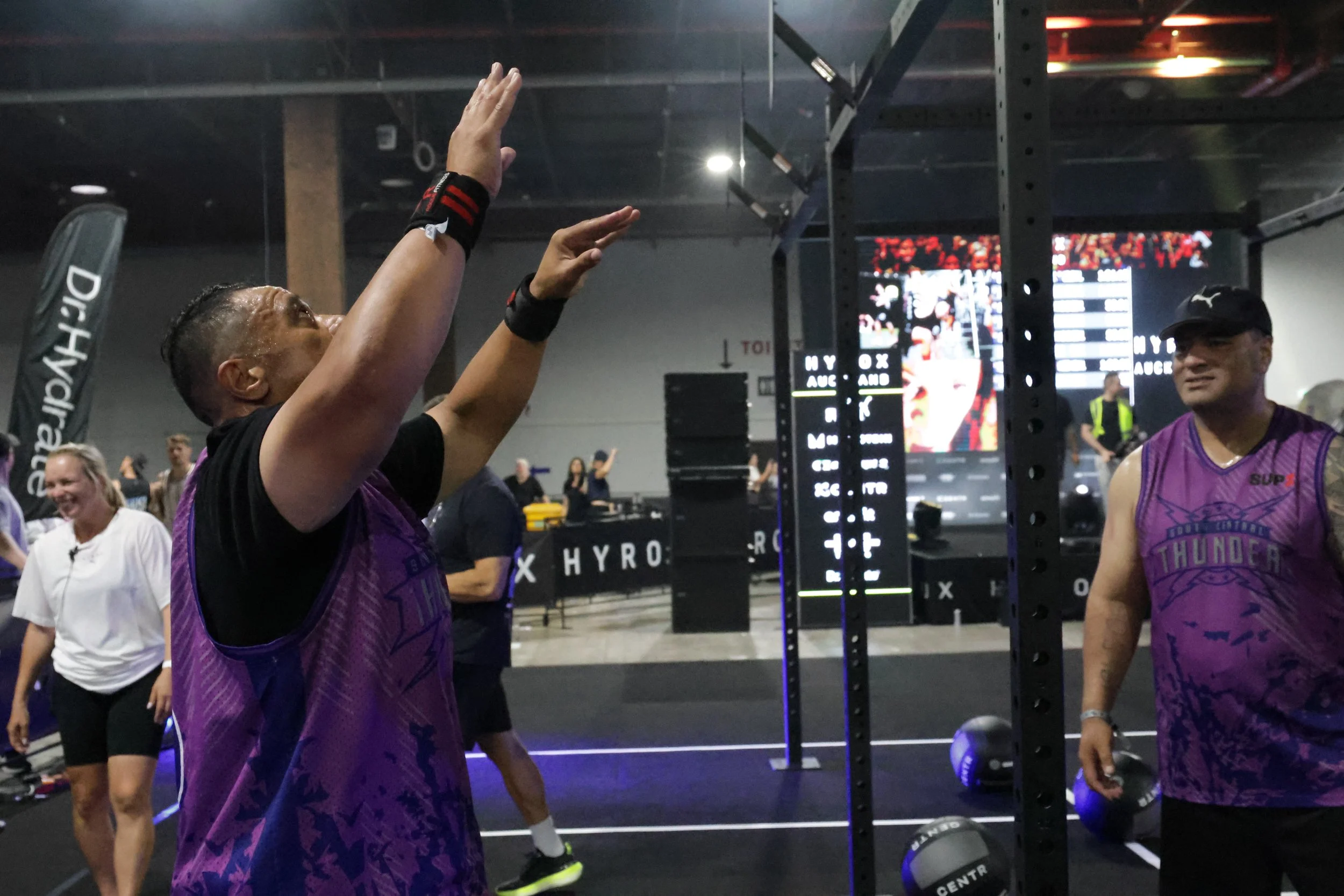 Two men in purple sports jerseys competing in a fitness competition, with one reaching towards a white ball and the other smiling, surrounded by other participants in an indoor gym with digital screens and equipment.