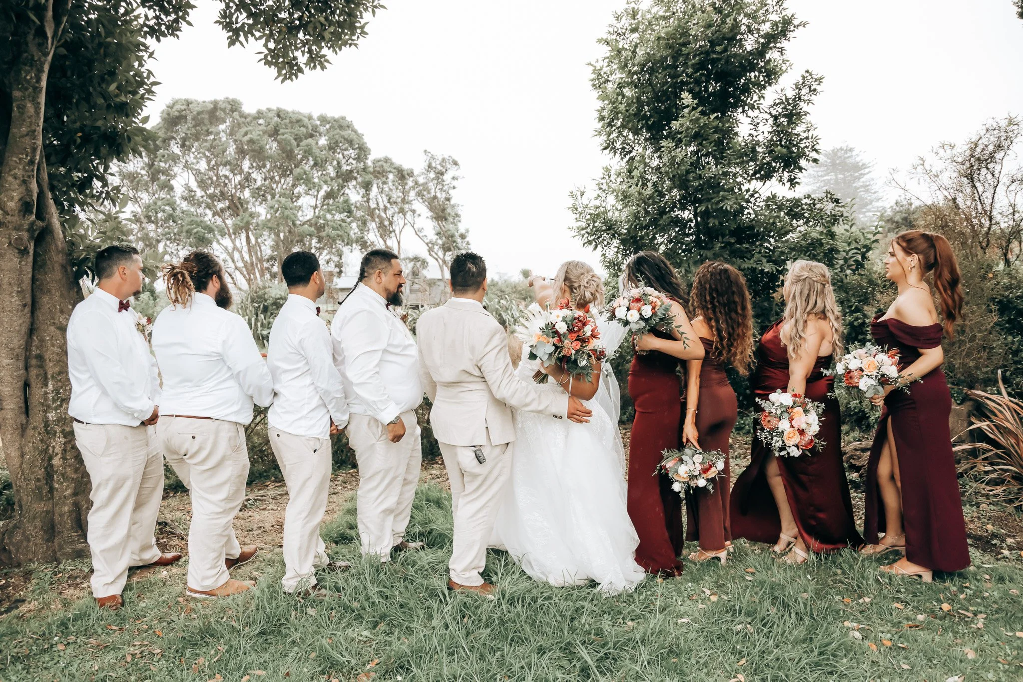 A wedding ceremony outdoors with the bride and groom holding hands, surrounded by bridesmaids and groomsmen. The group is standing on grass with trees in the background.