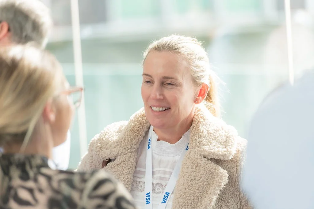 A woman with blonde hair smiling and talking to two other women in a bright indoor setting.