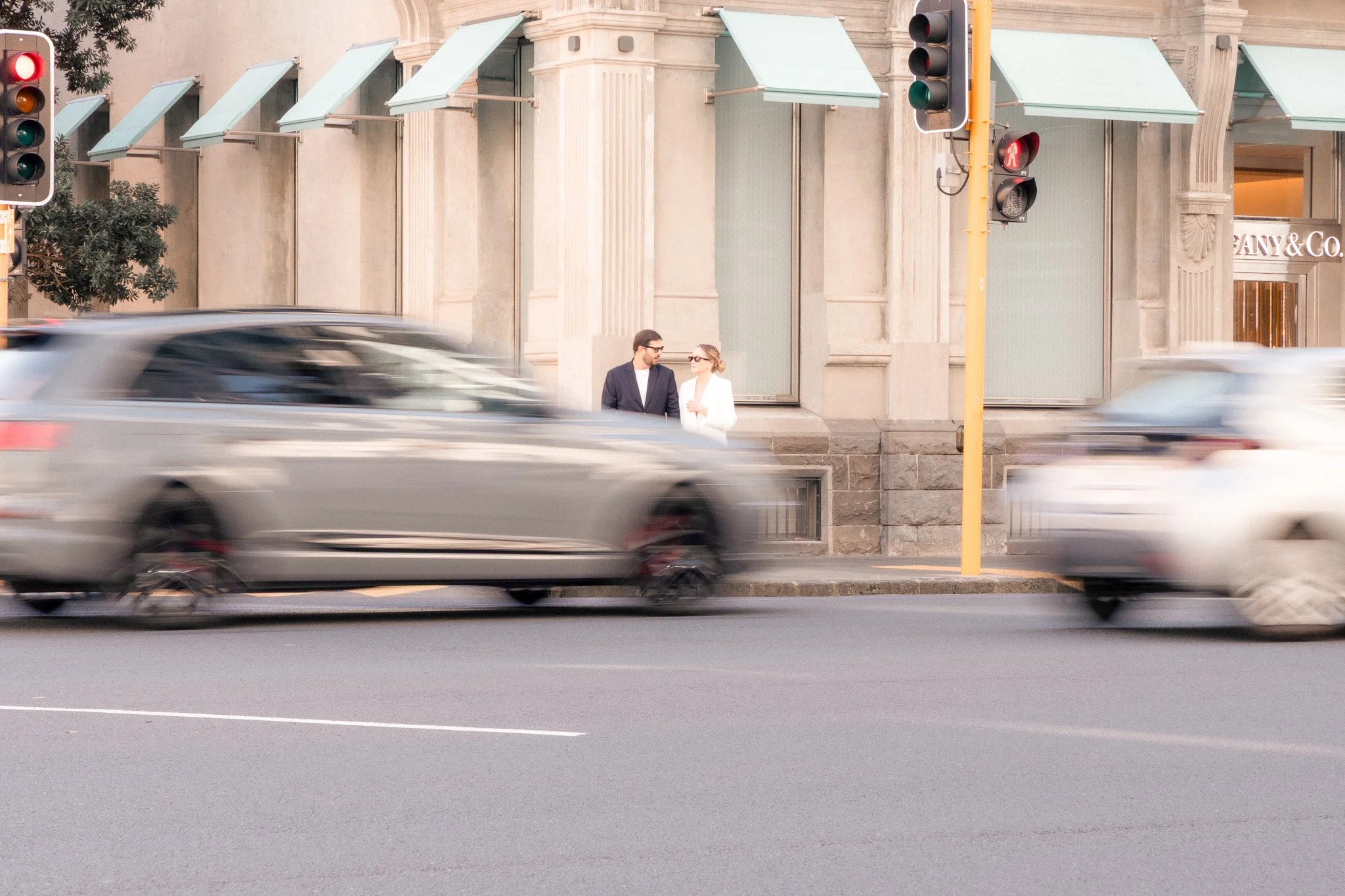 Street scene with two pedestrians standing on the sidewalk near a building, passing cars on the road, traffic lights, and storefronts.