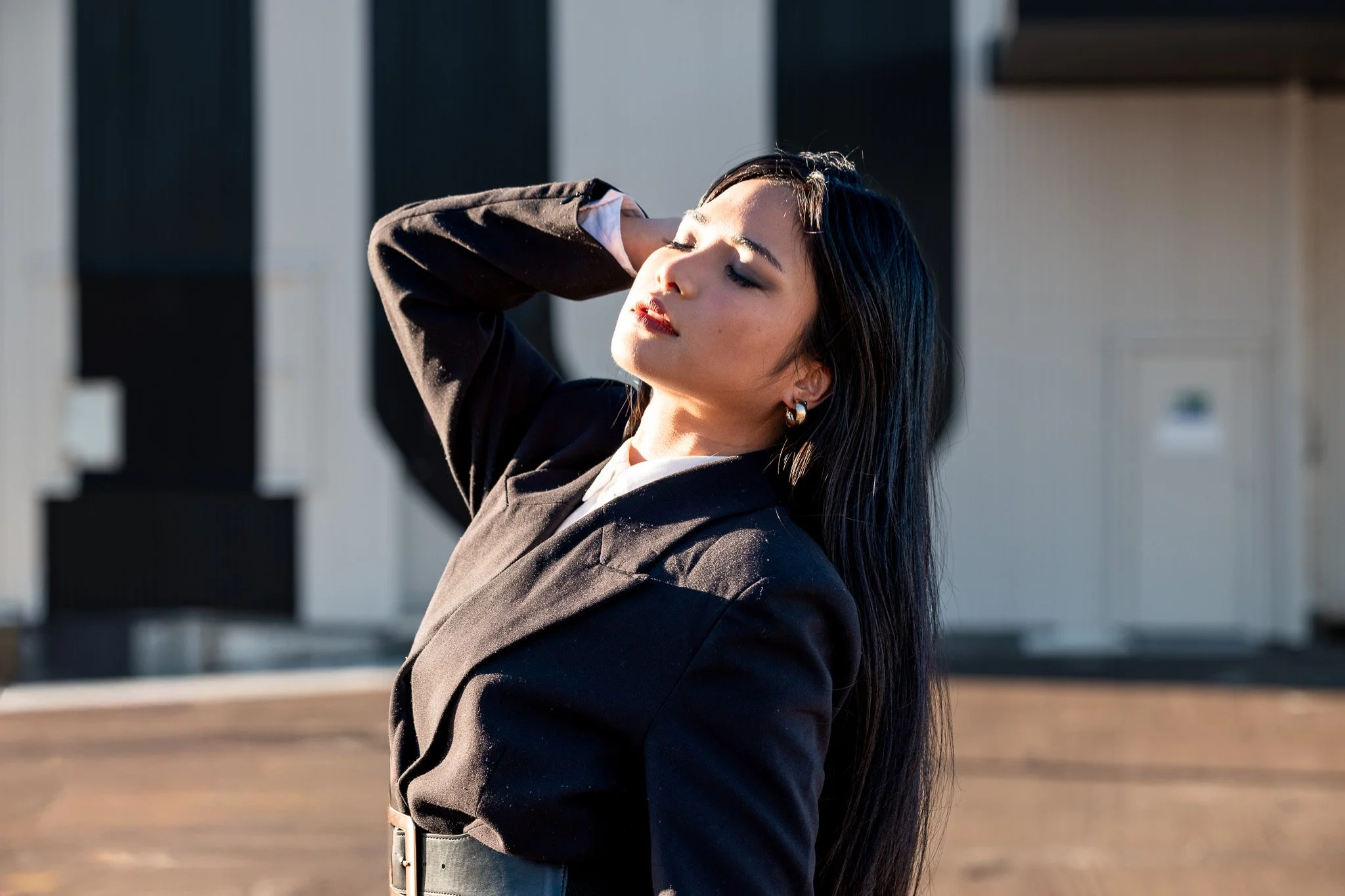 A woman with long dark hair and hoop earrings enjoys the sunlight with her eyes closed, standing outdoors against a background of modern black and white structures.