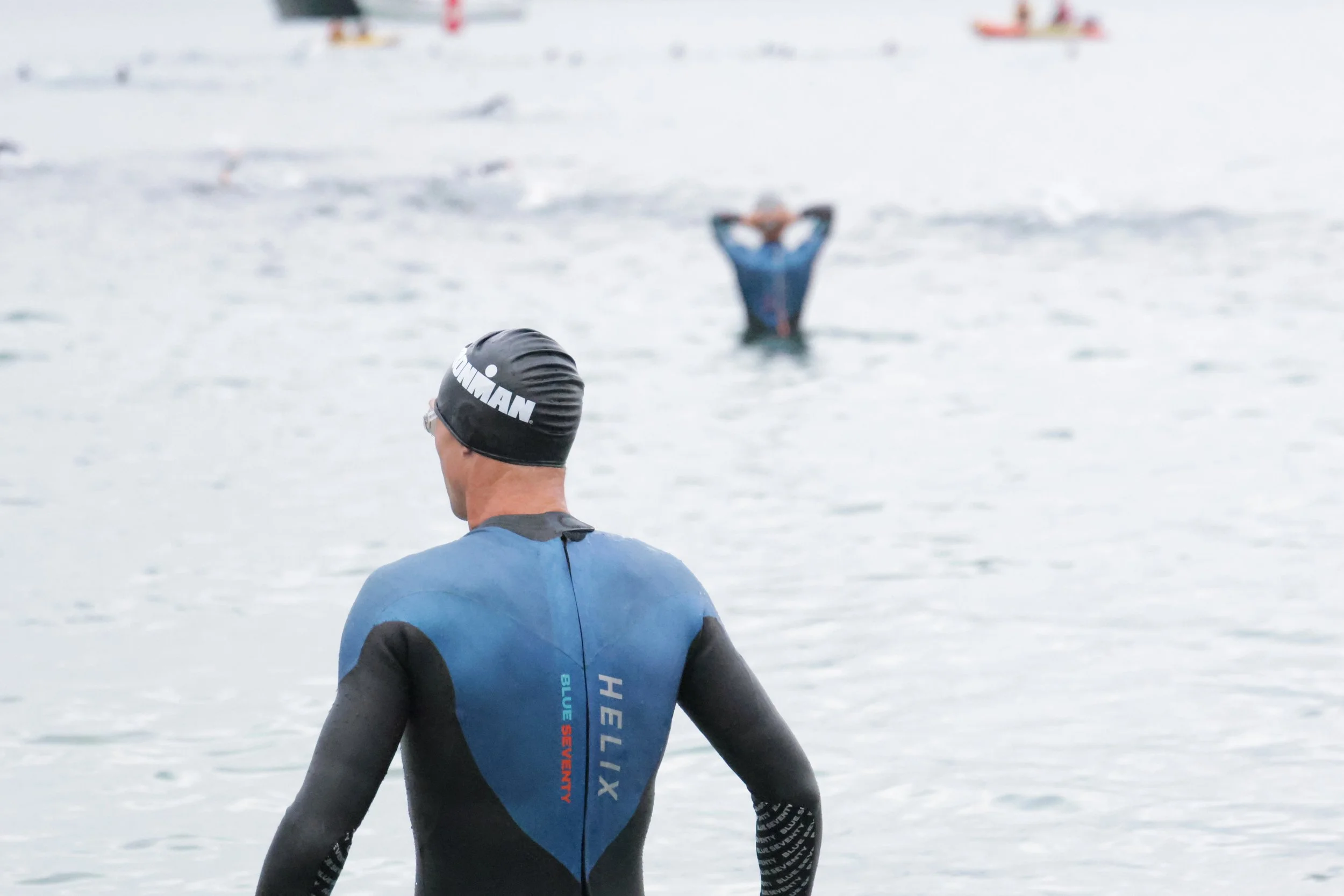 Swimmer wearing a black swim cap and wetsuit, standing in water with other swimmers in the background, blurred.