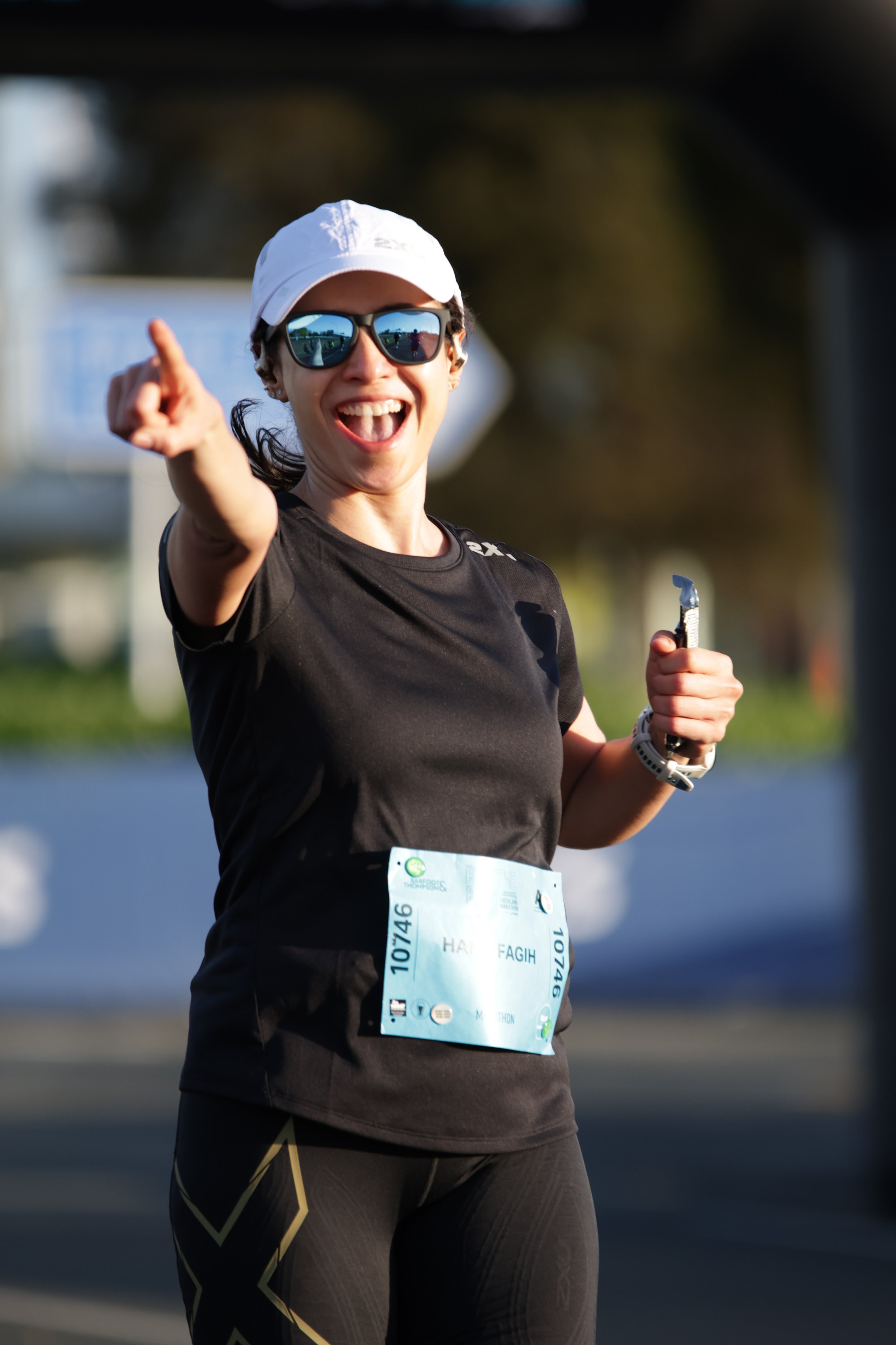 A smiling woman in athletic gear, sunglasses, and a white cap, pointing and holding a small object, participating in a race or marathon outdoors.