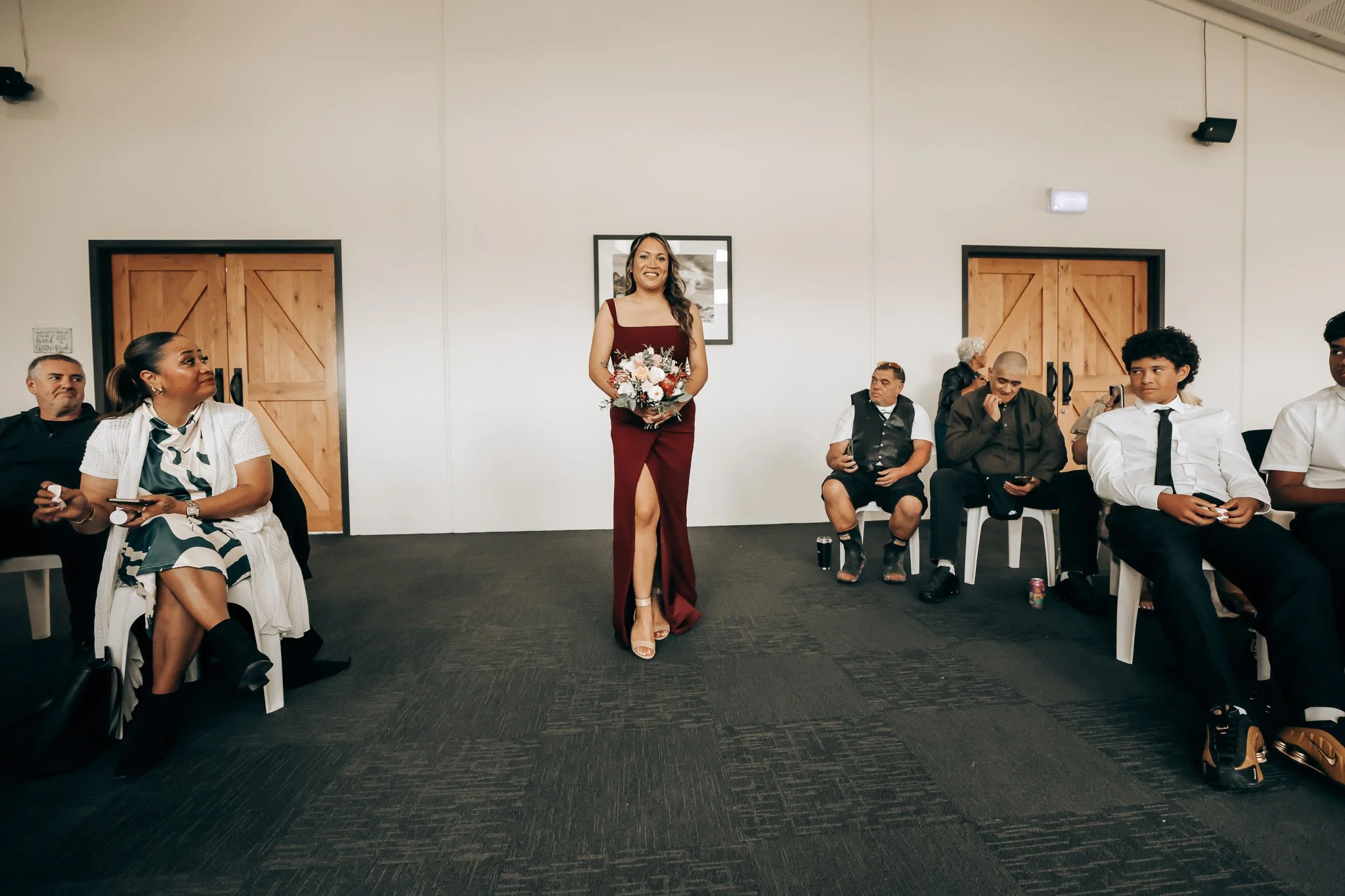 A woman in a burgundy dress holding a bouquet of flowers, standing in a room with several seated people watching her, some taking photos or videos.