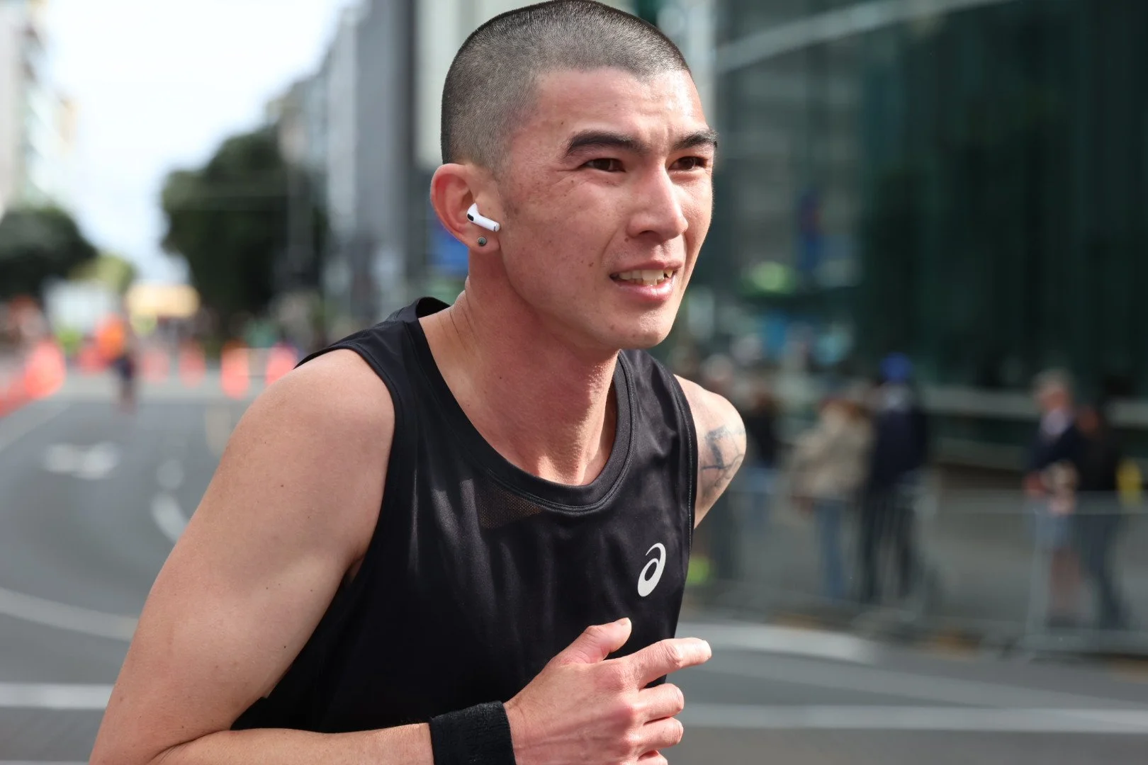 A male marathon runner with a shaved head, wearing a black running tank top and wireless earbuds, running outdoors in an urban setting during daytime.