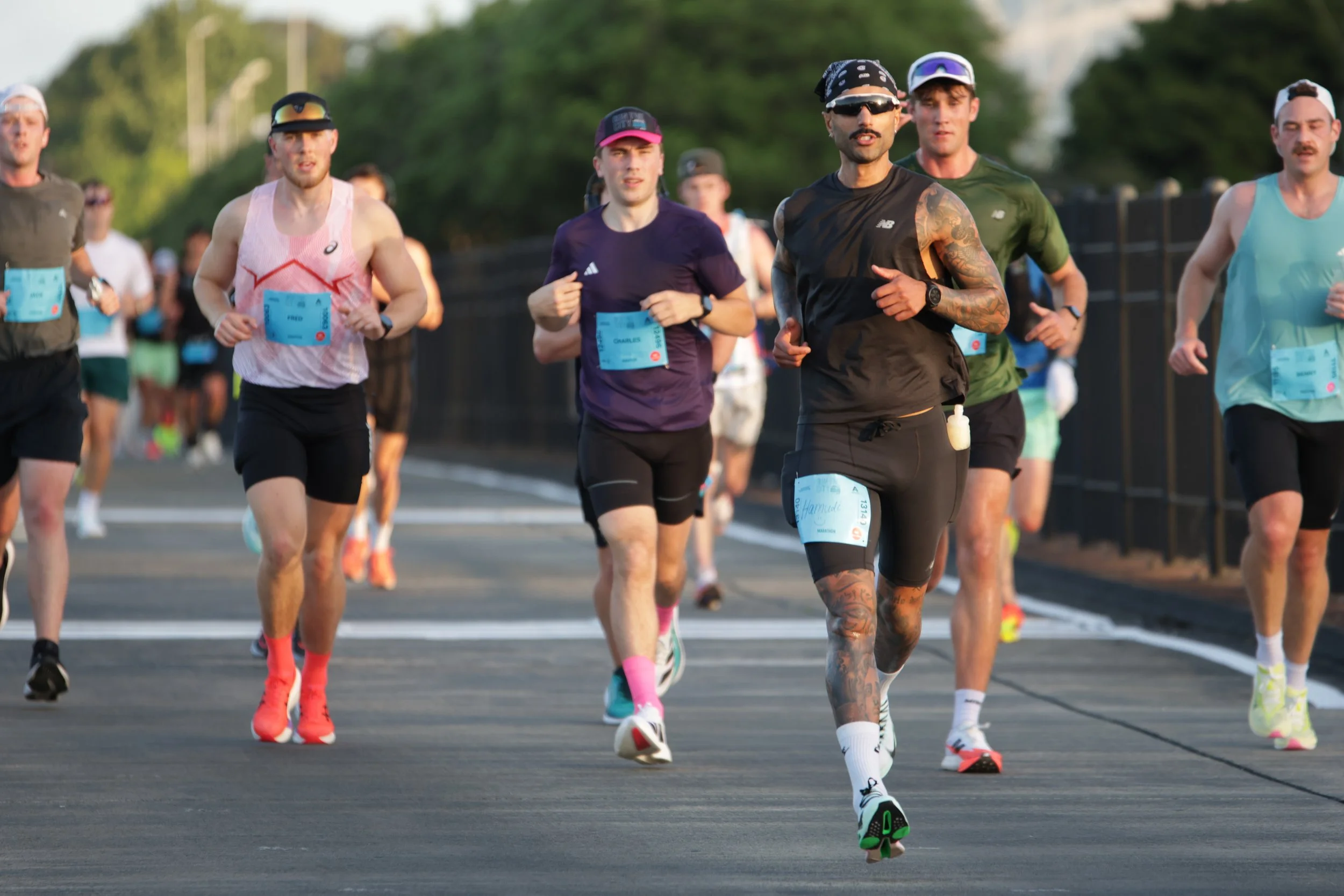 A group of runners participating in a race outdoors, wearing athletic clothing and race bibs, running along a paved road with trees in the background.