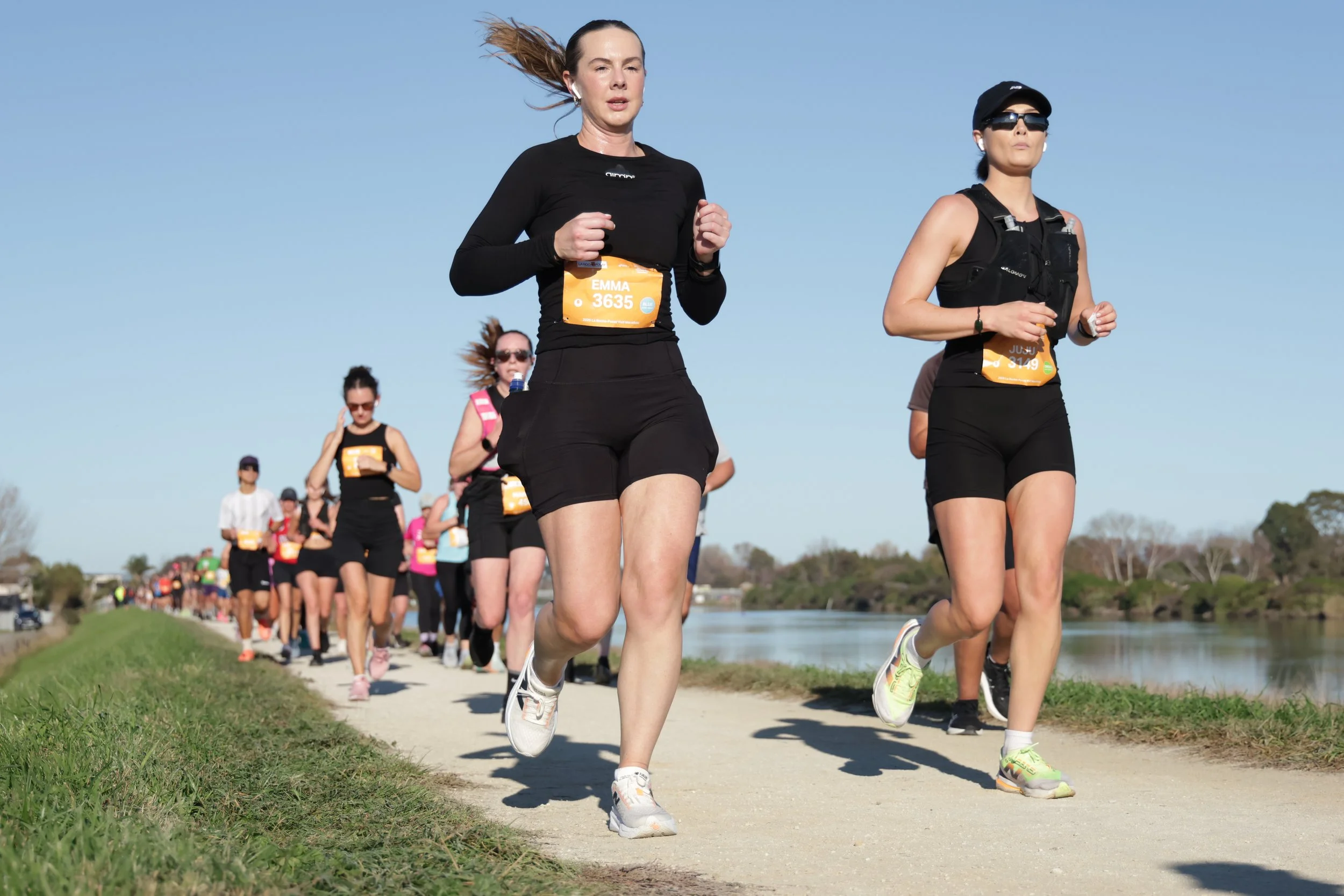 A group of women participating in a marathon or running event outdoors on a sunny day, running along a dirt path beside a body of water with trees in the background.