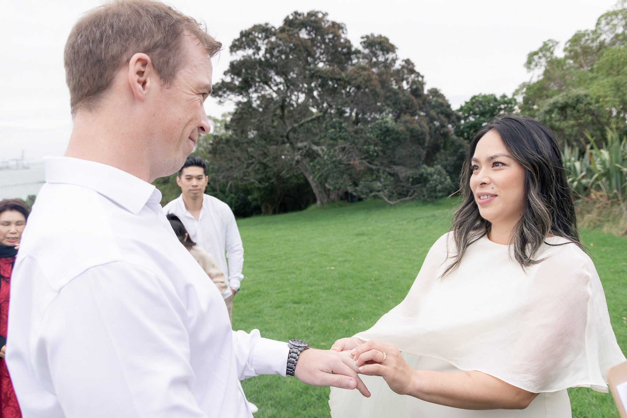 A man and a woman are holding hands and smiling at each other outdoors, with a group of people and trees in the background.