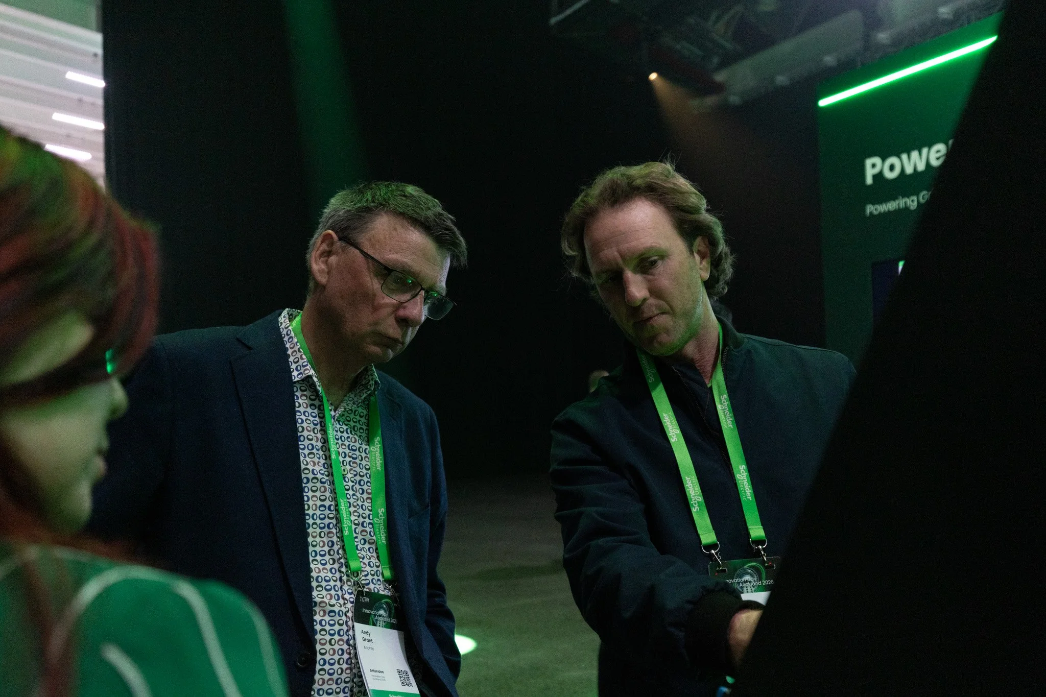 Two men with conference badges looking down at a device at an indoor event with dark lighting and green accents.