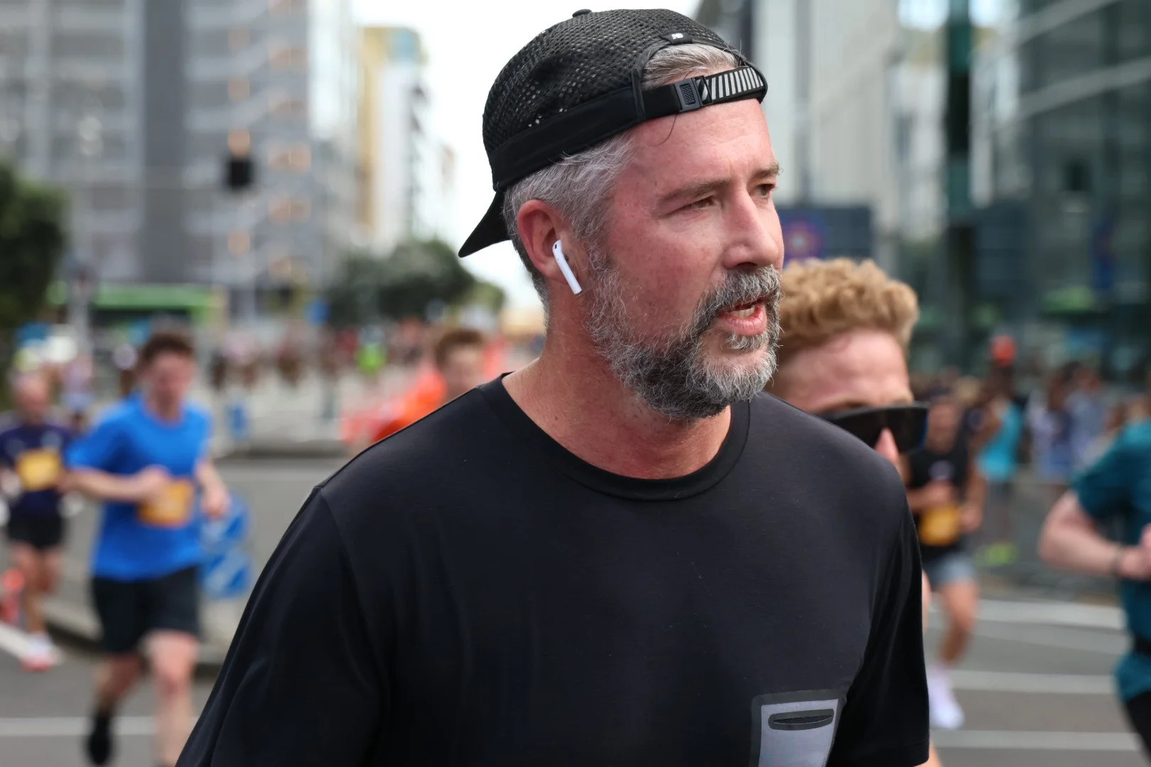 A man with gray hair and beard running in a marathon, wearing a black cap backwards, black shirt, and wireless earbuds, with city buildings in the background.
