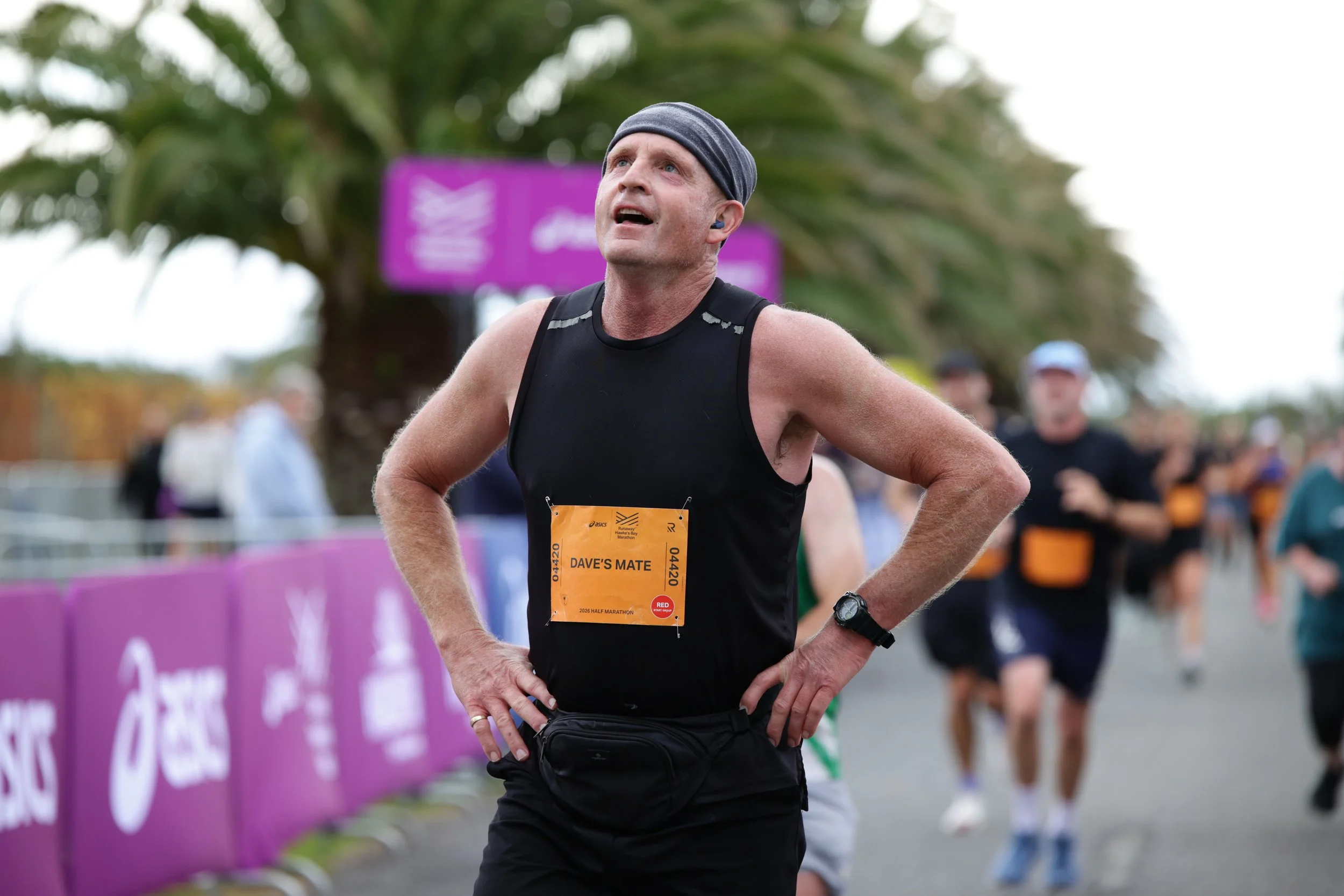 A middle-aged male runner with a race bib that reads 'DAVE'S MATE' stands with his hands on his hips, looking upward during a race. He is wearing a black tank top, a gray headband, and a watch on his left wrist. There are other runners and a purple b