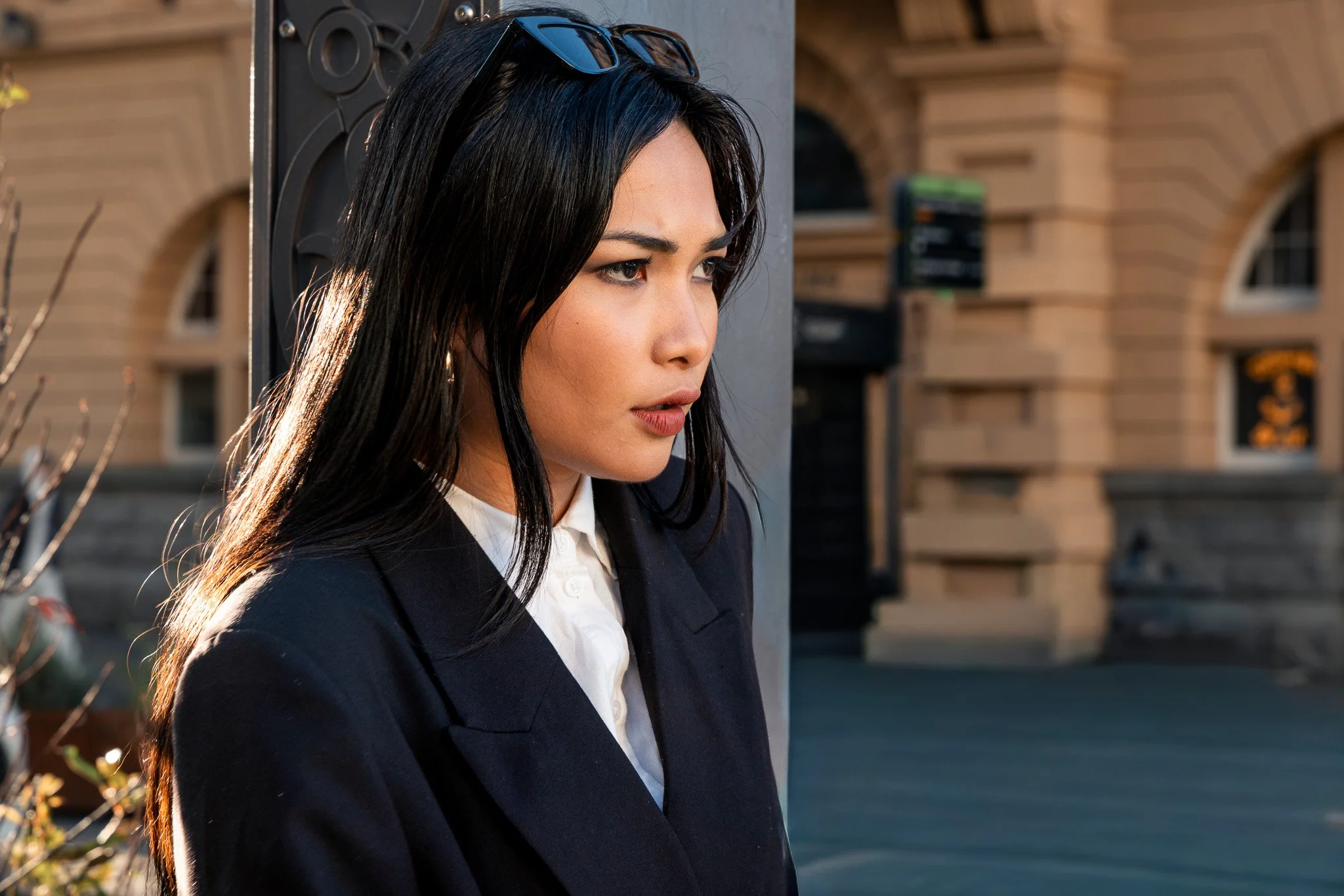 A young woman with long dark hair, wearing a black blazer and white shirt, standing outdoors in front of a building with arched windows, with sunglasses resting on her head and a serious expression.