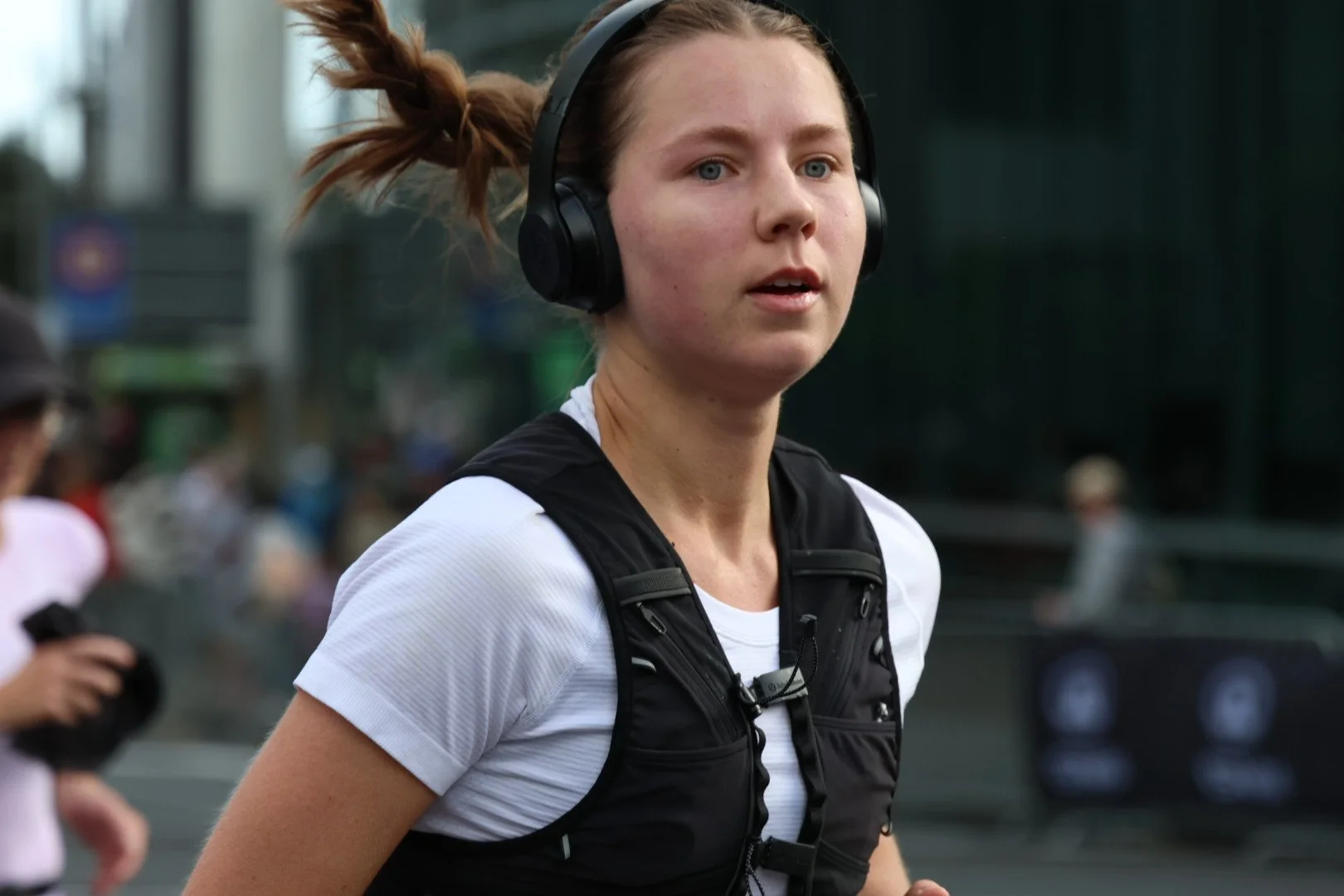 A young woman with red hair in a ponytail, wearing headphones and a white athletic shirt, runs outdoors in an urban setting during the day.