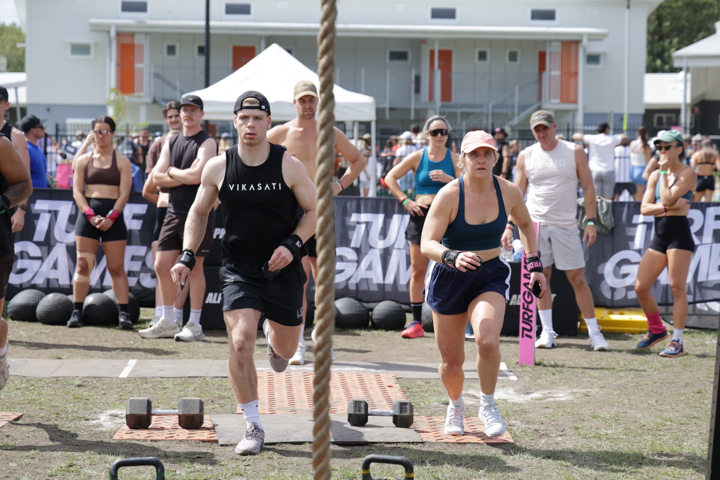 A group of people participating in an outdoor fitness event, with some walking or running, and others observing, in front of residential buildings and tents on a sunny day.
