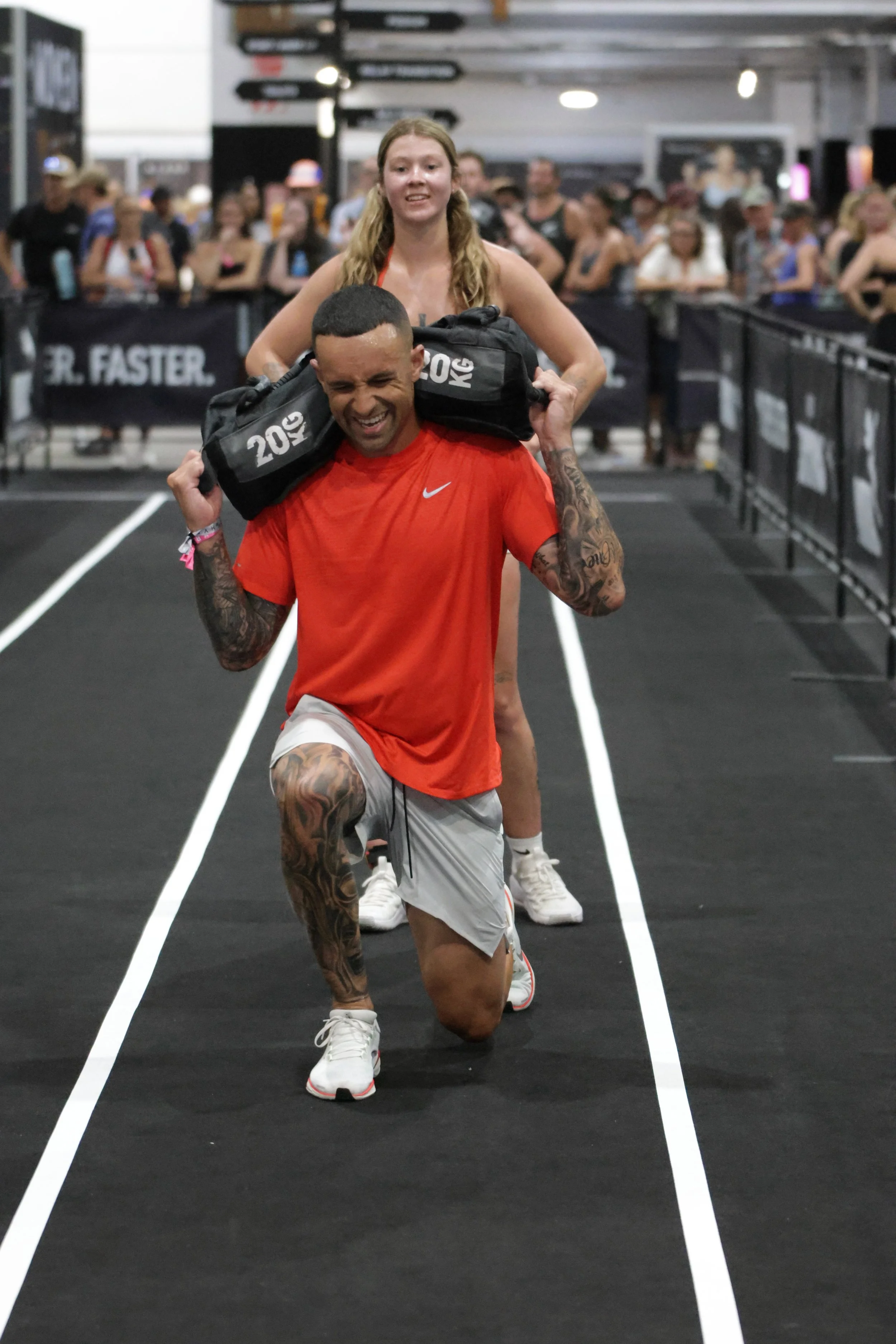 A man in a red shirt and gray shorts is running on an indoor track while carrying a black sandbag on his shoulders. Behind him, a woman with long blonde hair in a black sports bra and shorts is smiling. There is a crowd of people in the background wa