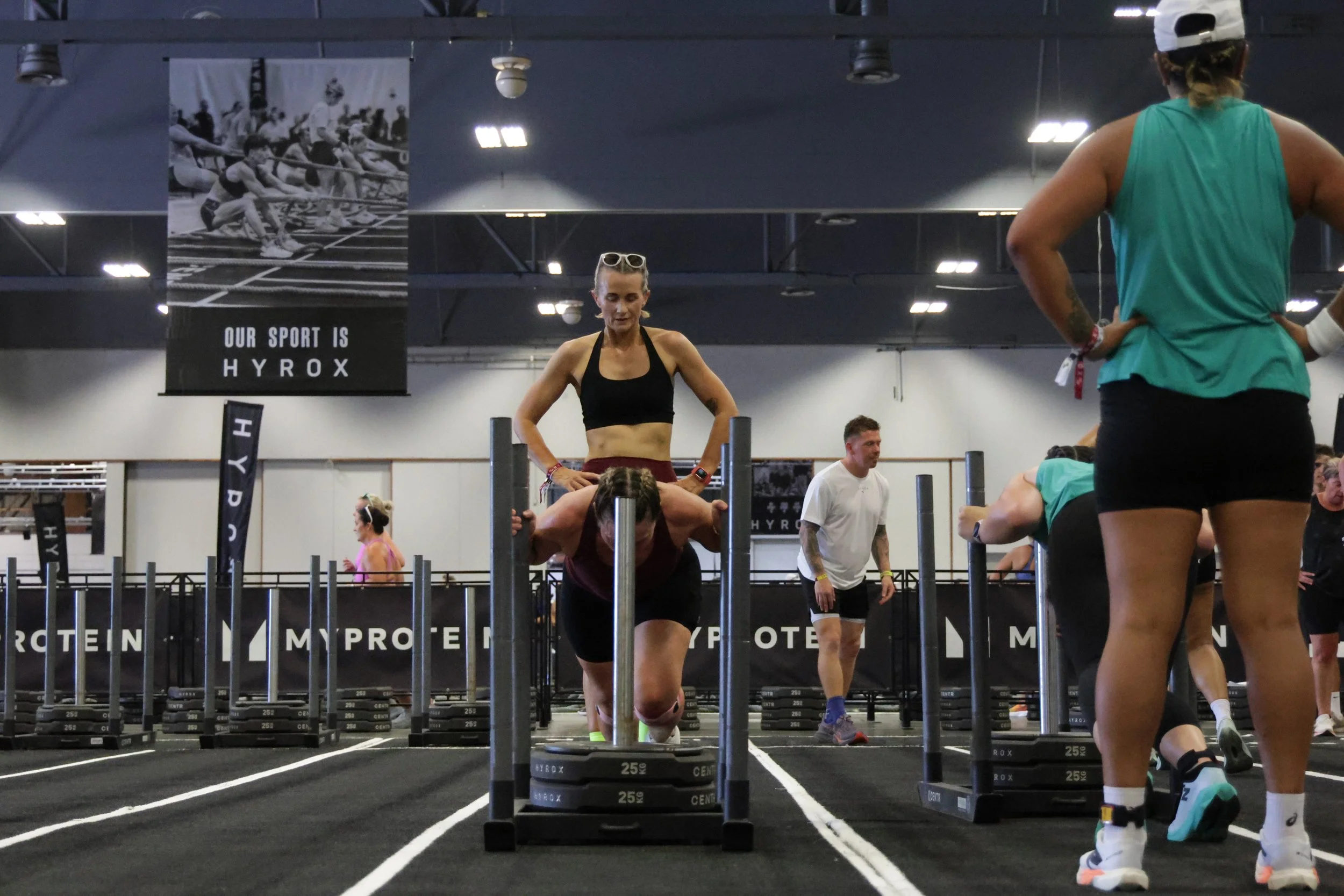 People exercising in a gym with a woman standing on a workout platform and others stretching or resting nearby.