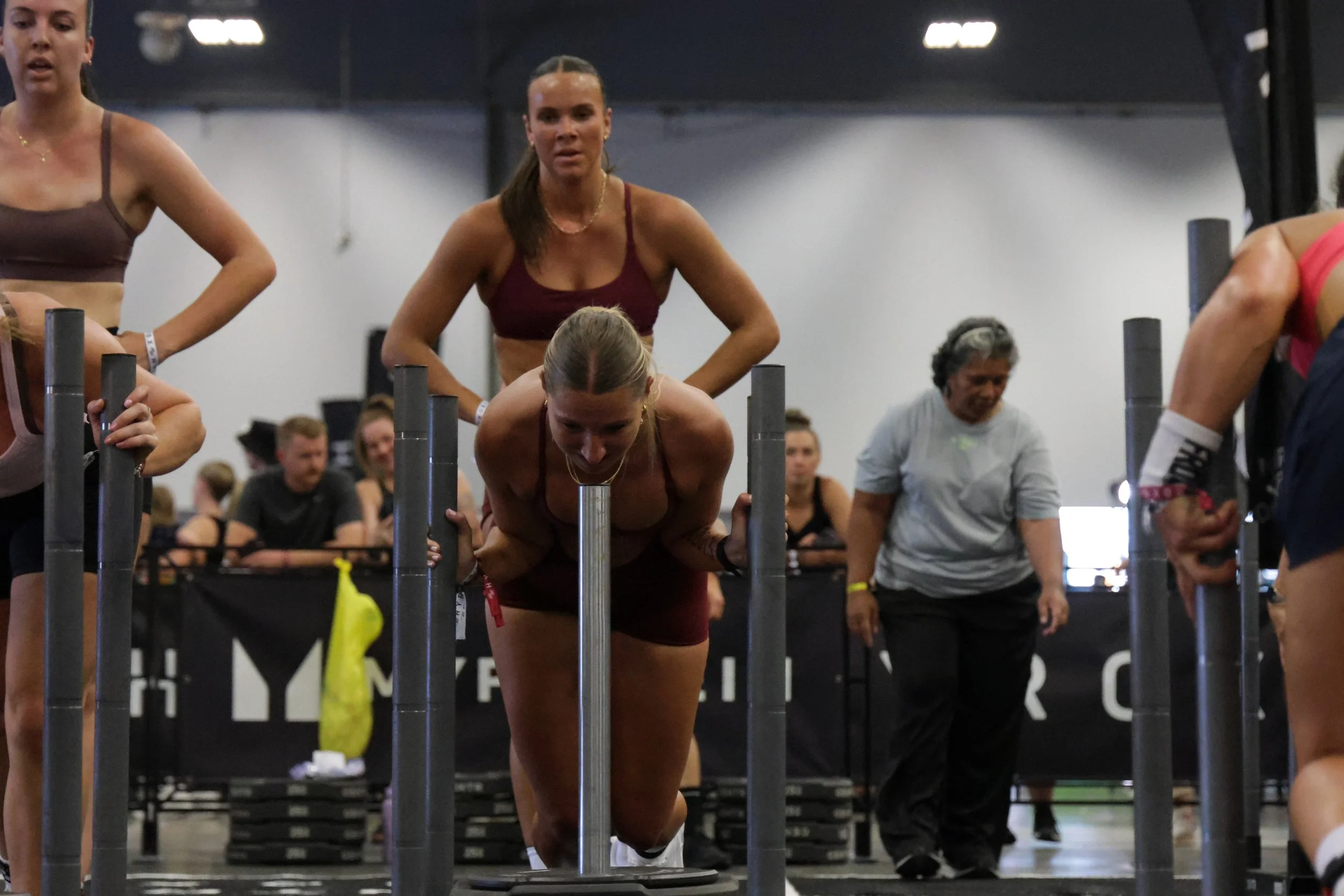 Women participating in a fitness or CrossFit event, with some performing push-ups and others watching, in an indoor gym setting.