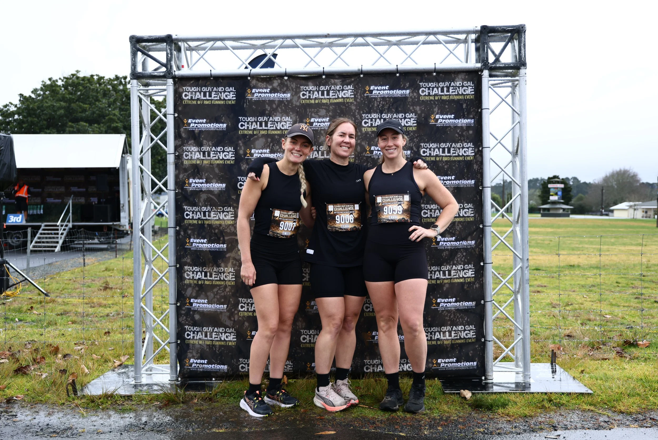 Three women in athletic gear standing together in front of a race event backdrop, smiling at the camera.
