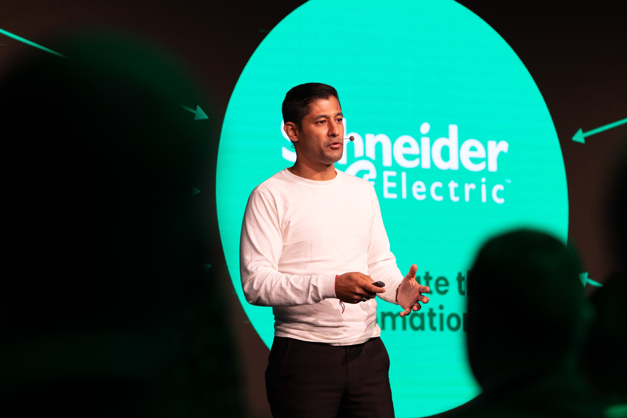 A man in a white shirt speaking at a presentation with a microphone headworn, standing in front of a large digital screen displaying the words 'Shneider Electric' in white text on a large blue-green circle, with arrows pointing towards it.