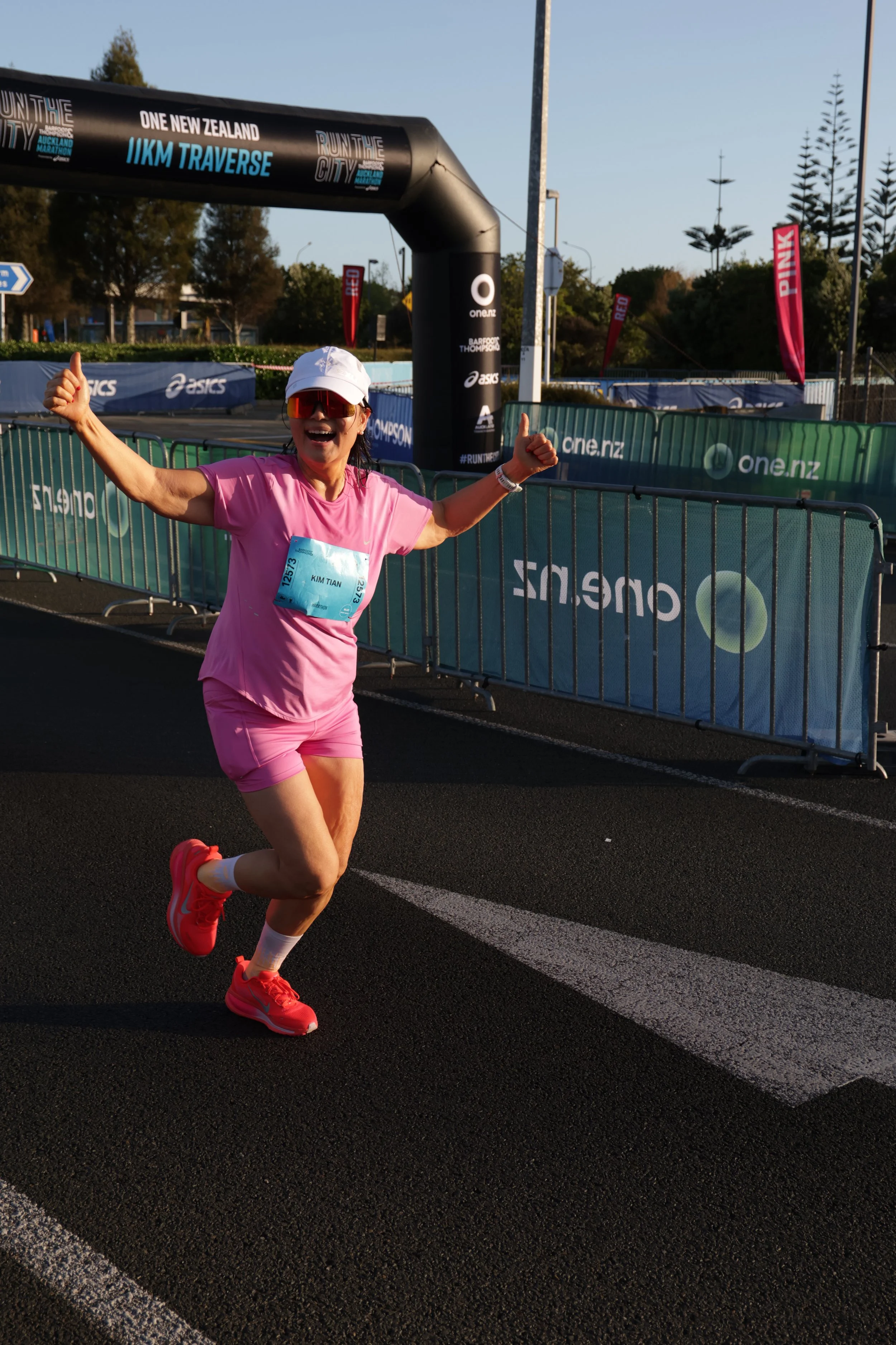 A woman running and smiling at the finish line of a marathon, wearing a pink shirt and shorts, white cap, red sunglasses, and pink running shoes near a fenced course with banners and an arch overhead that reads 'Run the City, IIKM Traverse, One New Z